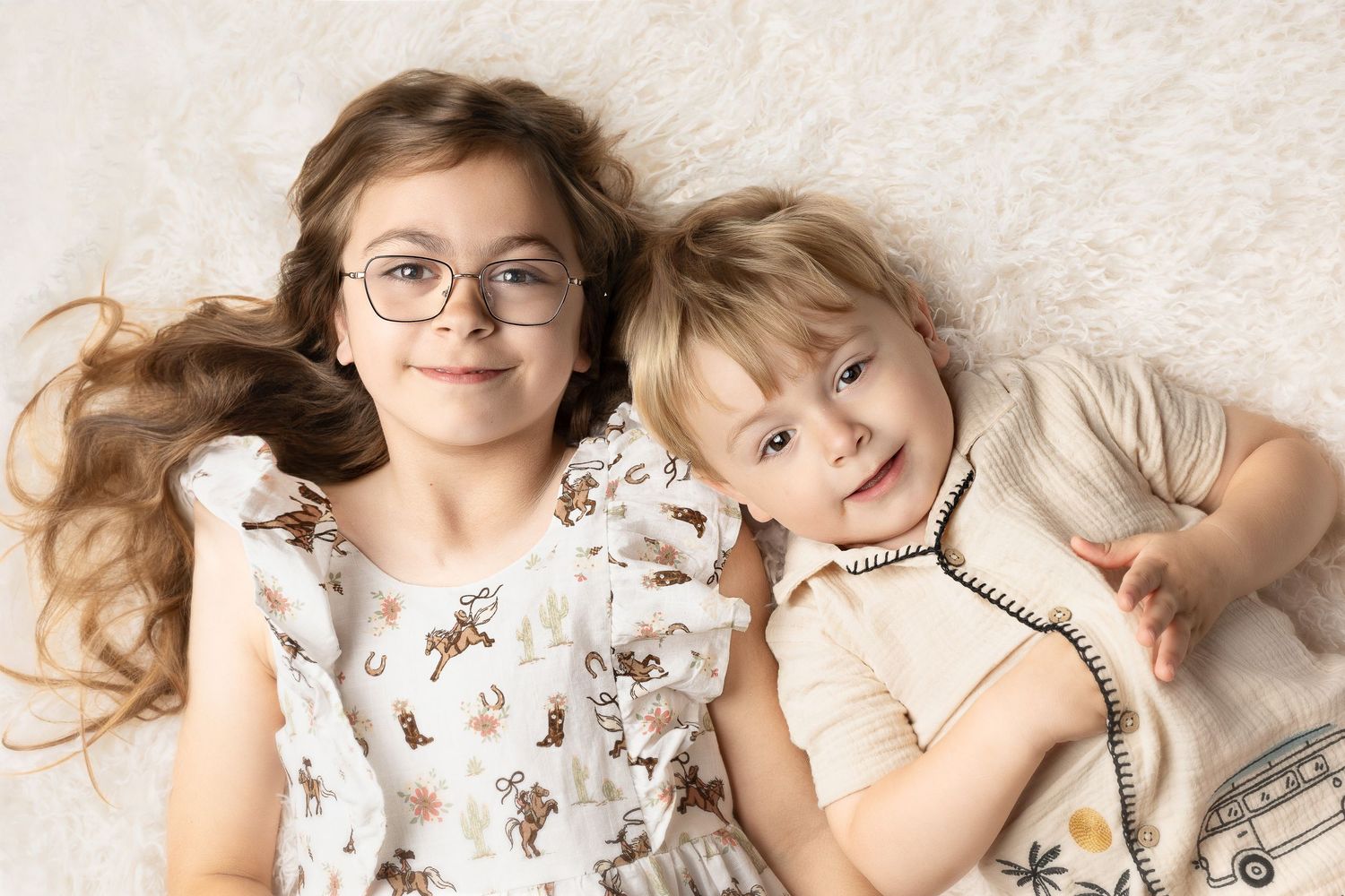 Two children lying on a white fluffy rug, a girl with glasses and long hair beside a younger boy.