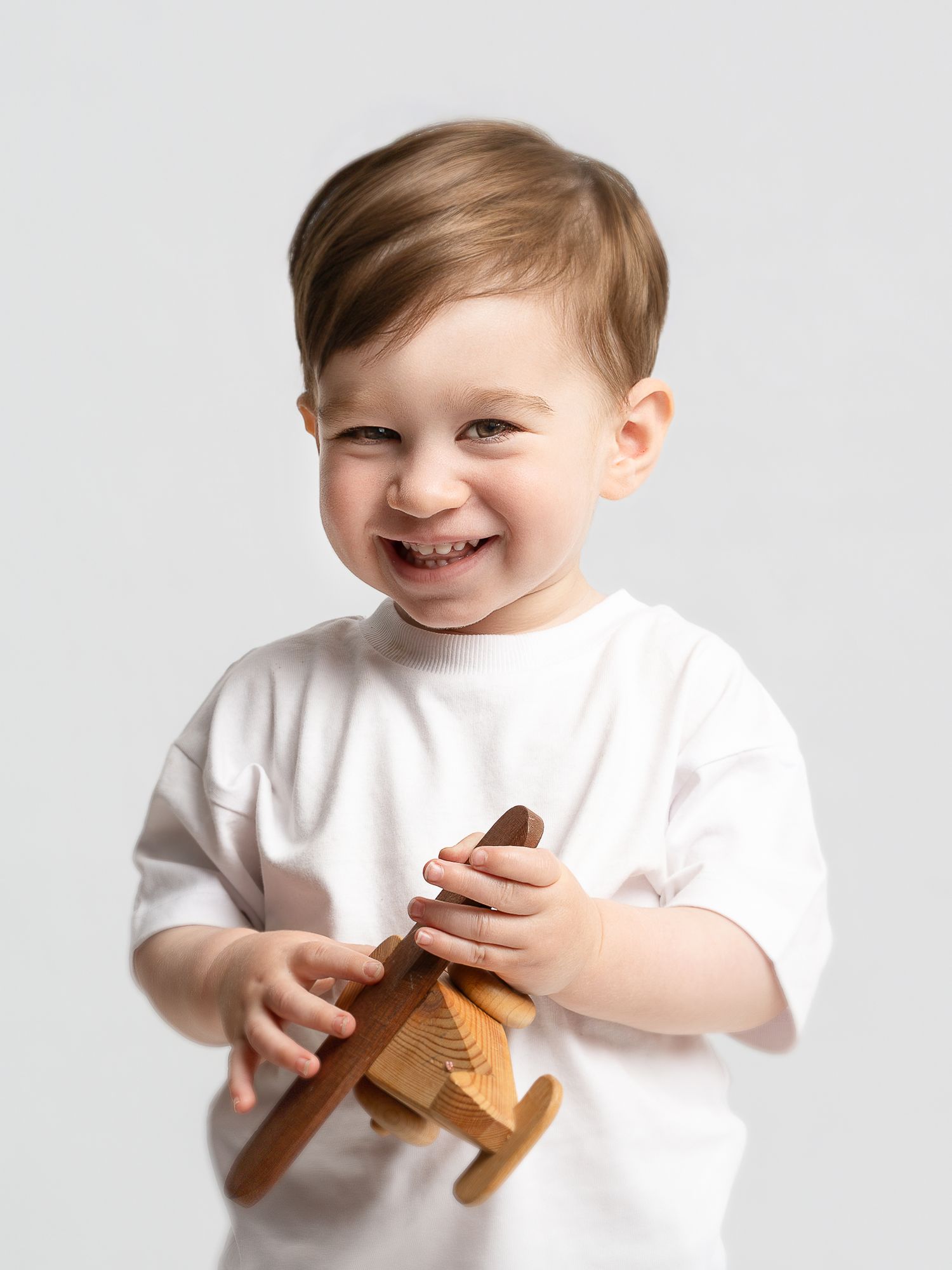 Smiling toddler in white t-shirt holding a wooden toy airplane against a light gray background.