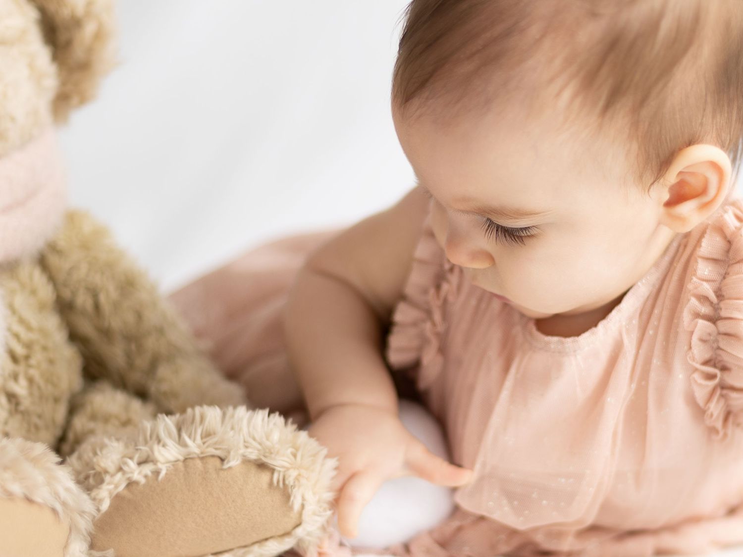 A baby girl in a pink ruffled dress gently touches a soft teddy bear during a professional newborn photo session.