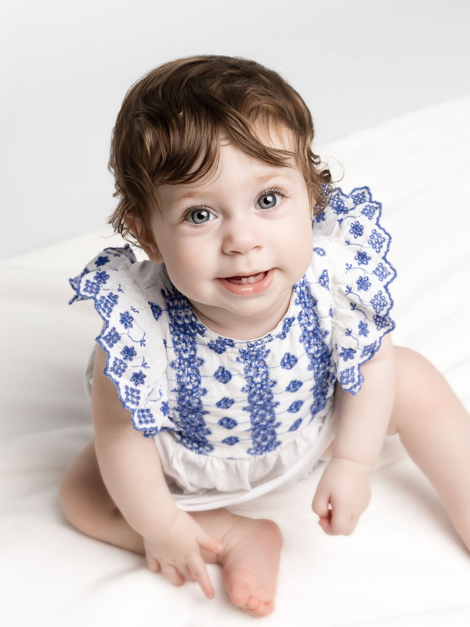Adorable baby girl in white and blue embroidered romper sitting up and smiling at camera.