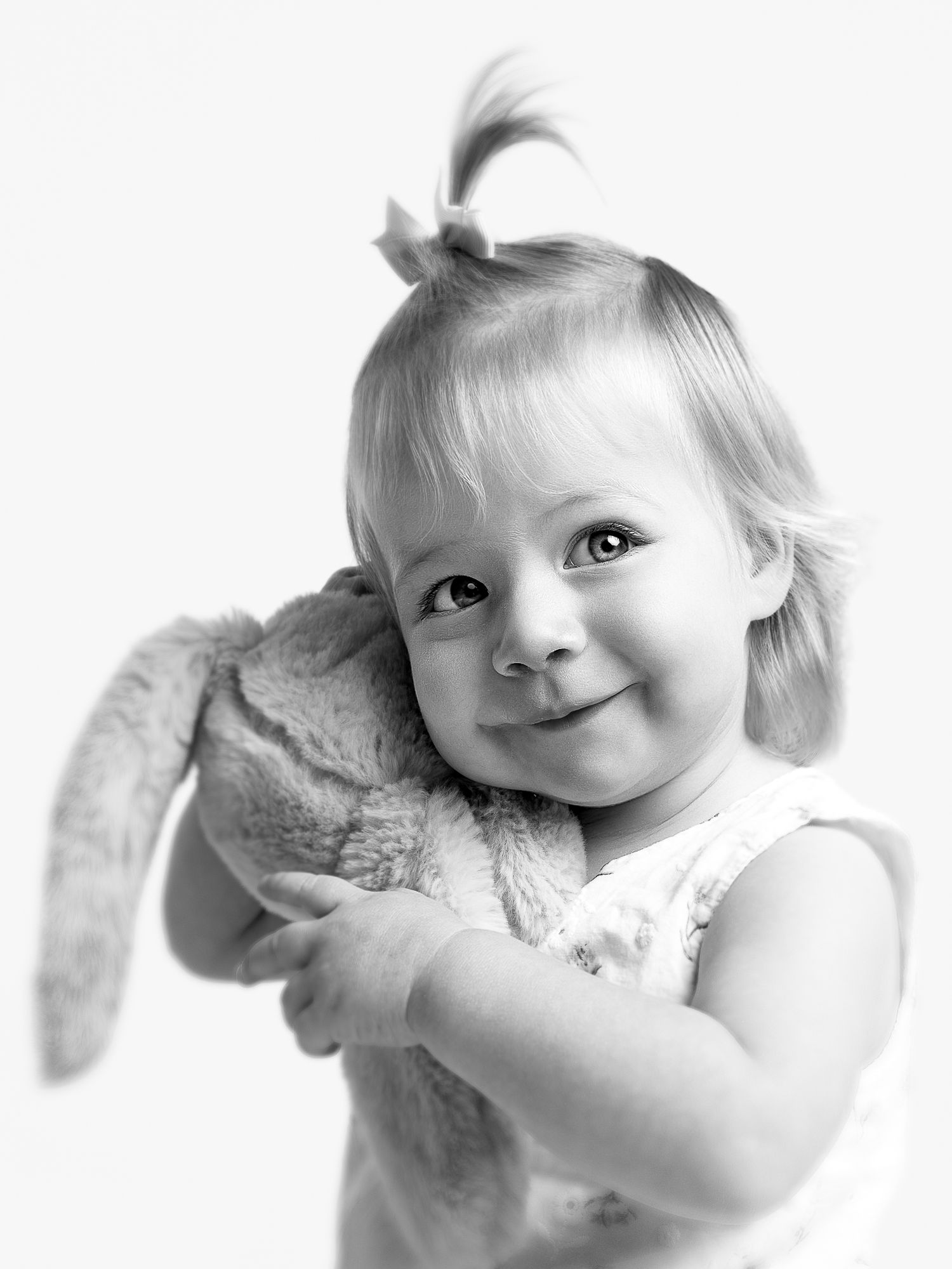 Black and white portrait of a smiling toddler girl hugging a stuffed bunny toy, with a small hair bow.