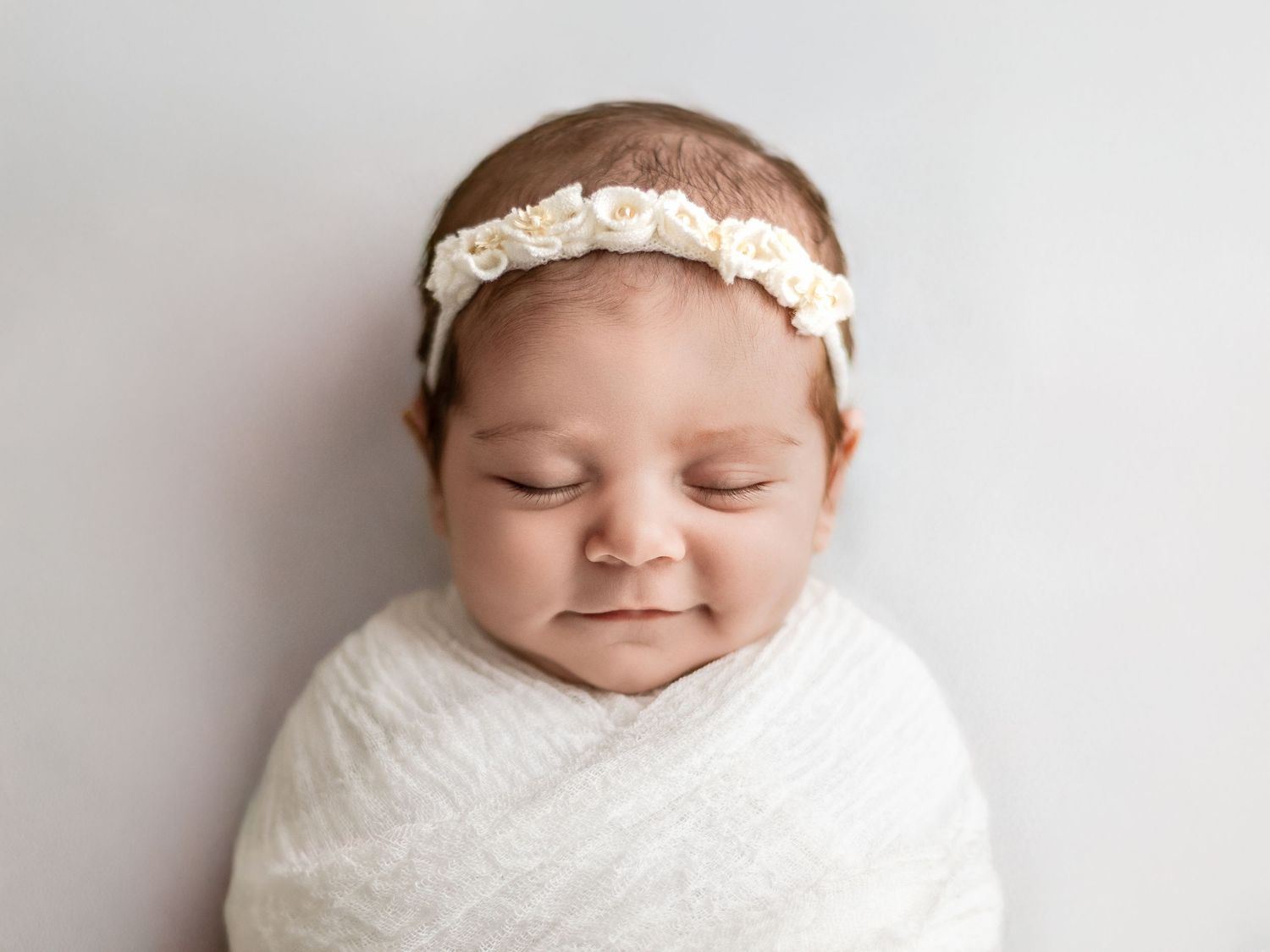 Newborn baby wearing white headband wrapped in white blanket against light background.