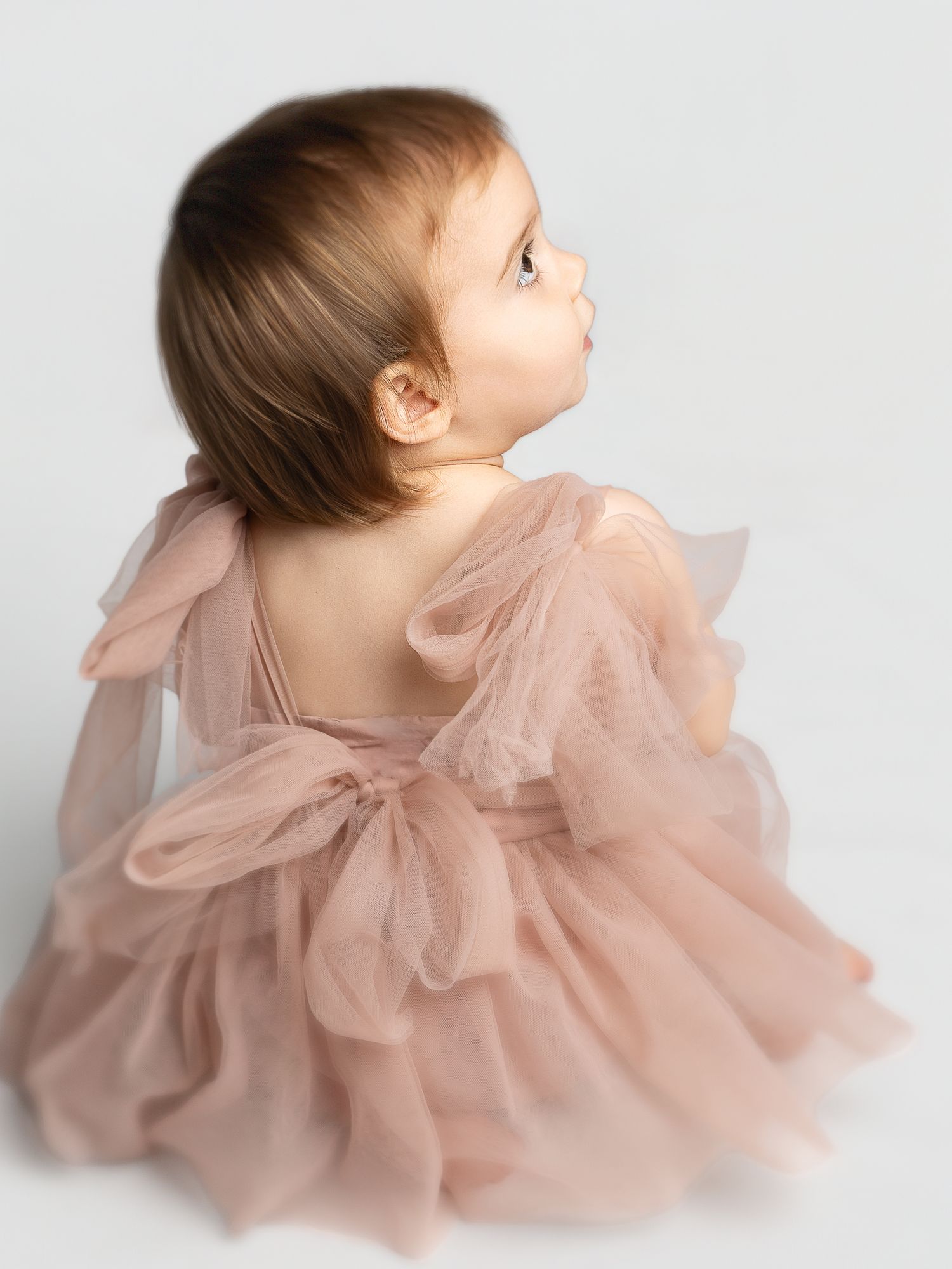 Baby girl in a blush pink tulle dress with bow details, photographed from behind on a white background.