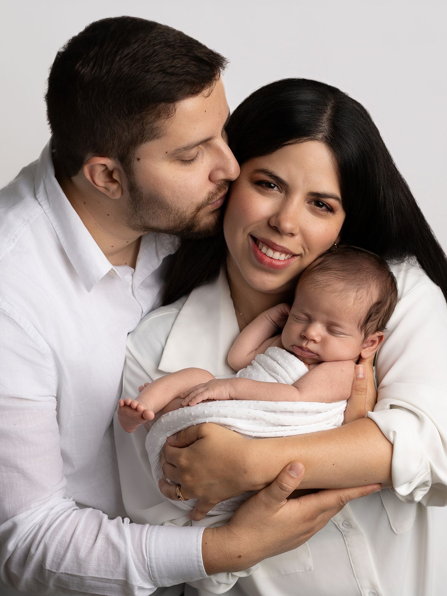 A couple in white shirts tenderly holds their sleeping newborn baby in a professional studio portrait photo.