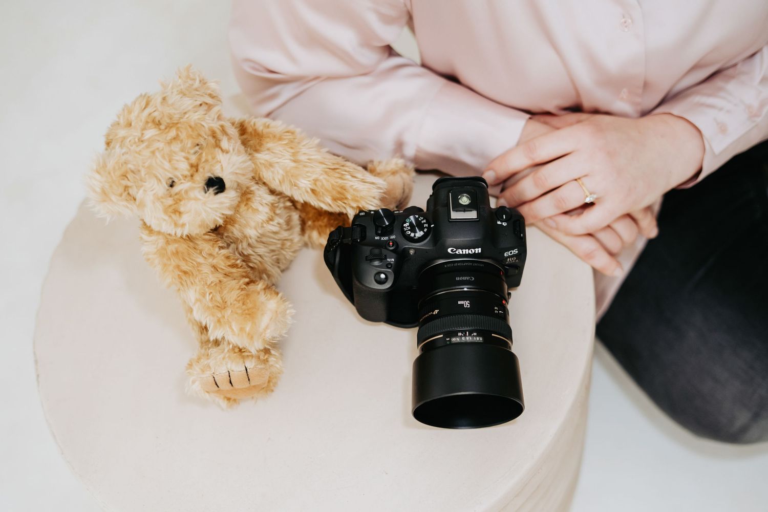 Person holding a Canon DSLR camera beside a fluffy teddy bear on a white table in a bright studio setting.