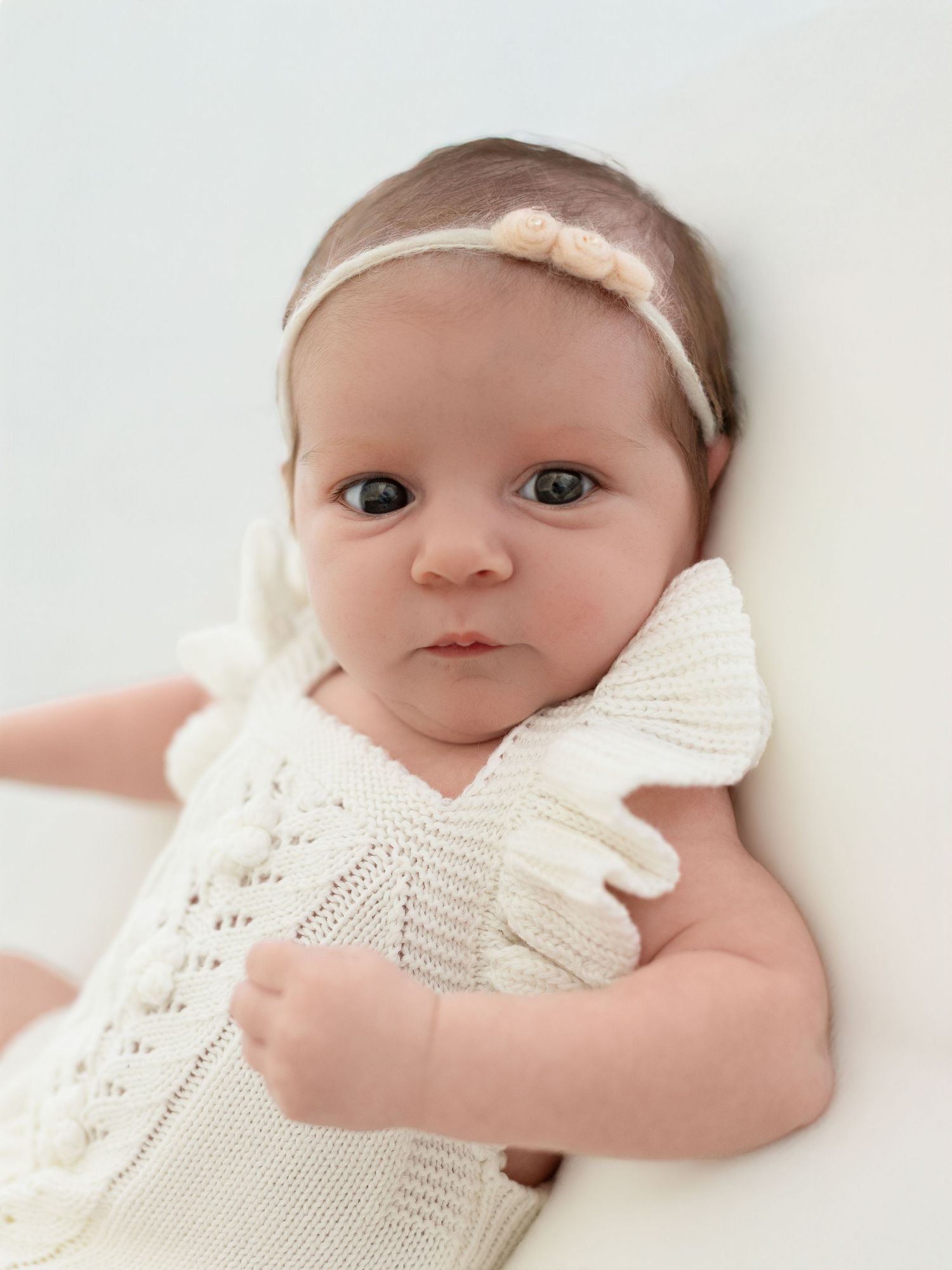 A newborn baby girl in a white knit romper and floral headband gazes at the camera in a soft portrait.