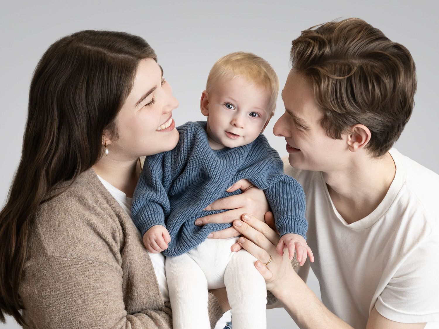 Young parents smiling at their blonde baby dressed in a blue knit sweater against a light gray background.