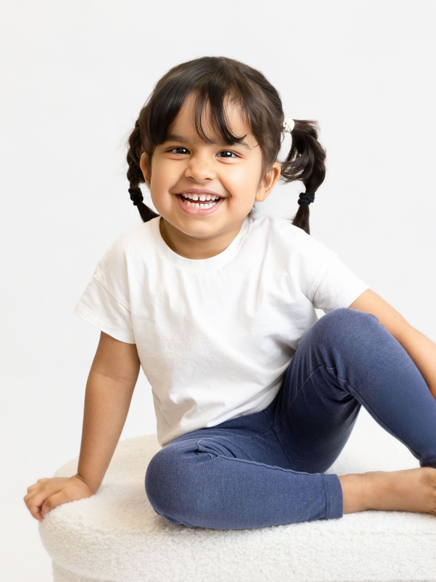 Smiling toddler girl with pigtails wearing a white t-shirt and blue leggings, sitting on a white pouf.