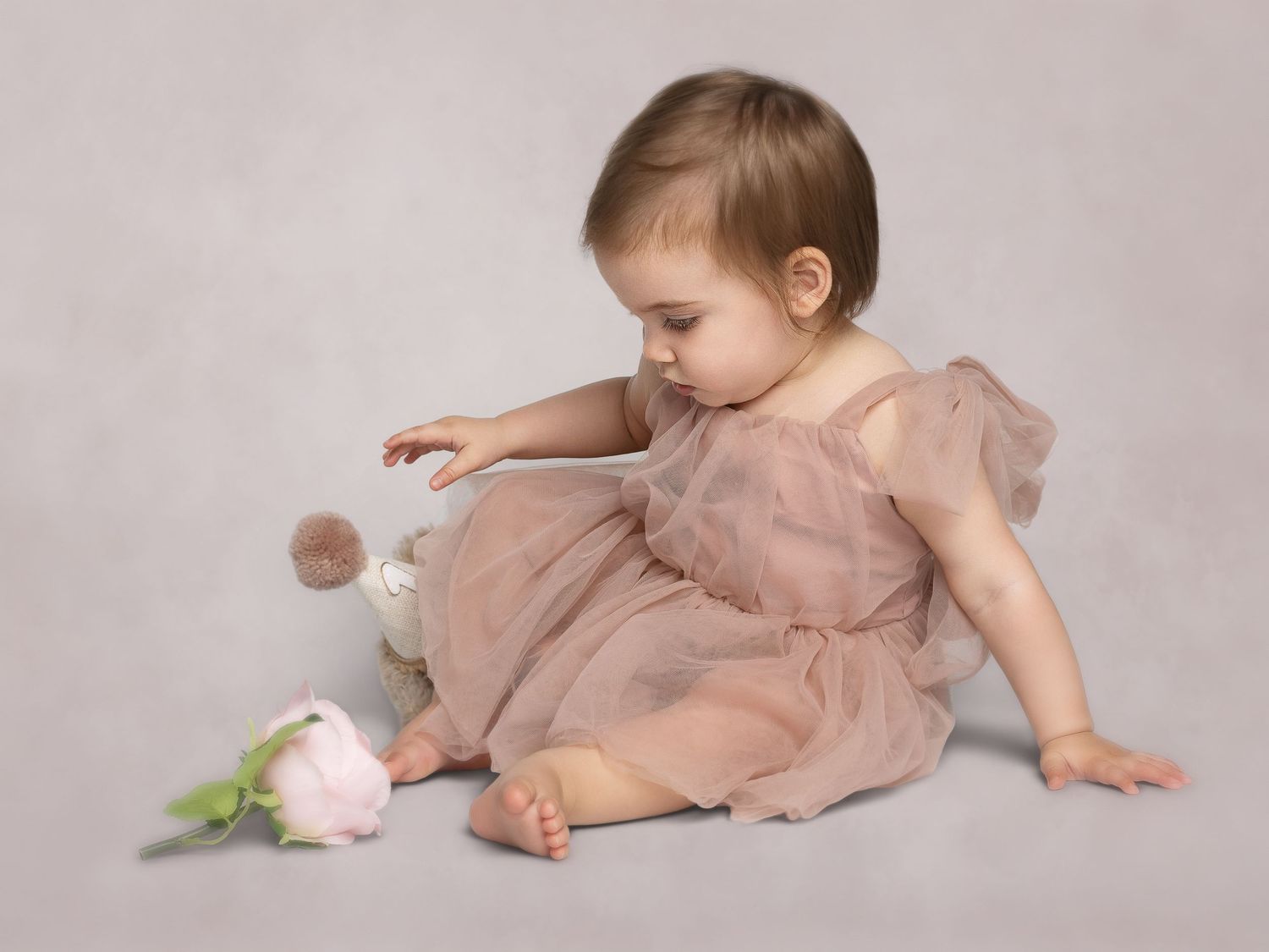 A baby girl in a pink tulle dress sits on a studio floor, gently touching a soft bunny toy beside a pink flower.