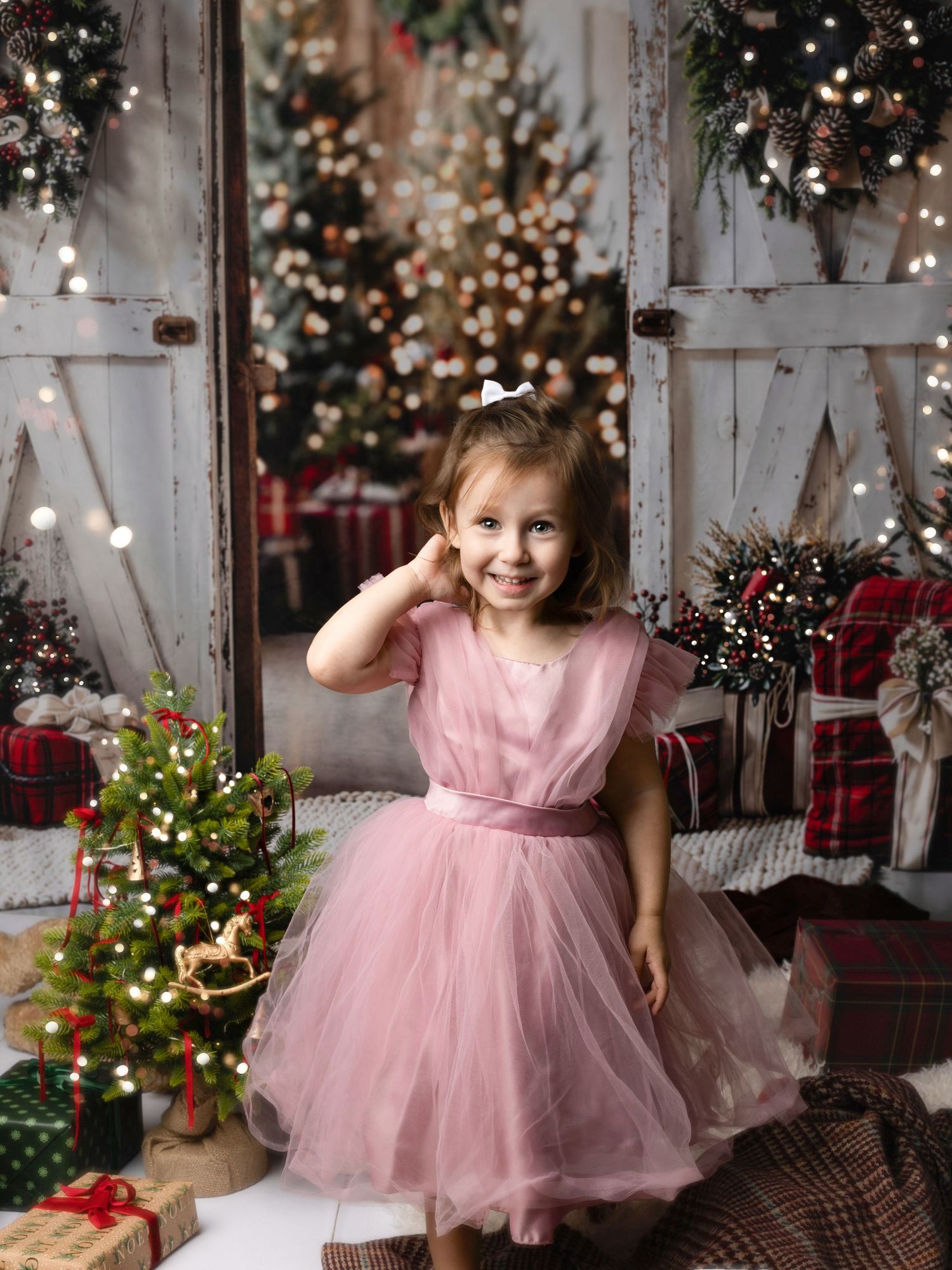 A smiling little girl in a pink tulle dress poses in a festive Christmas setting with decorated trees and gifts.
