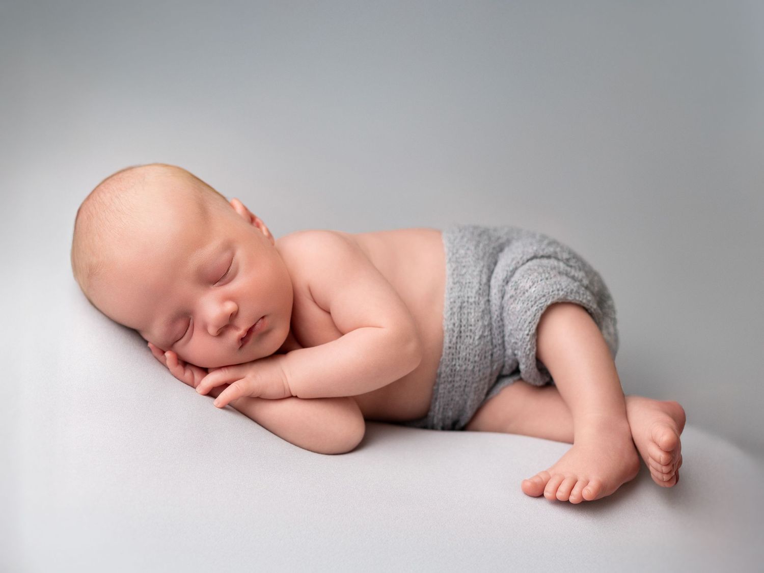 Side view of sleeping newborn baby wearing grey wrap on light backdrop.