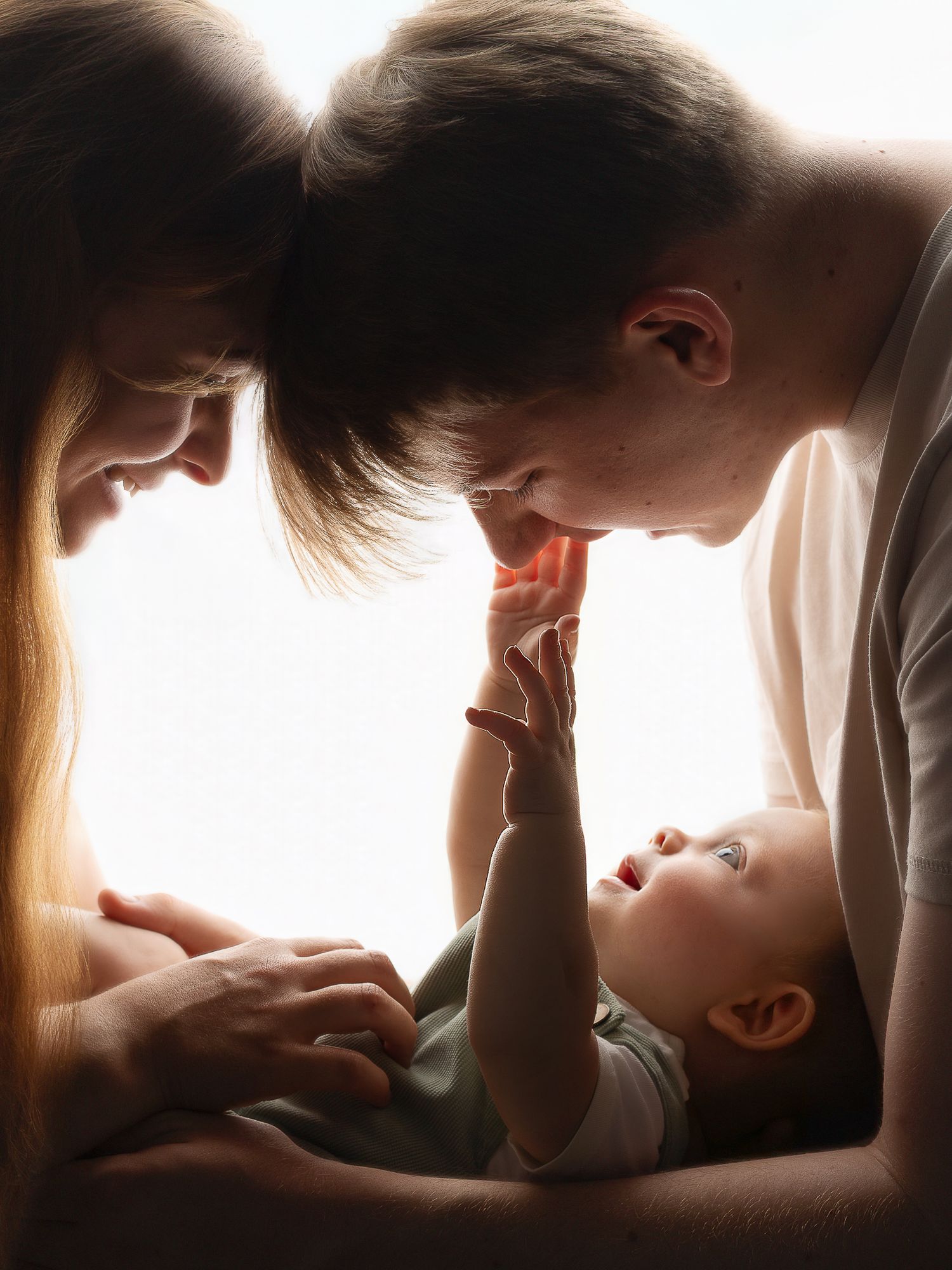 Two parents leaning over their smiling baby, sharing a tender family moment in soft backlit light.