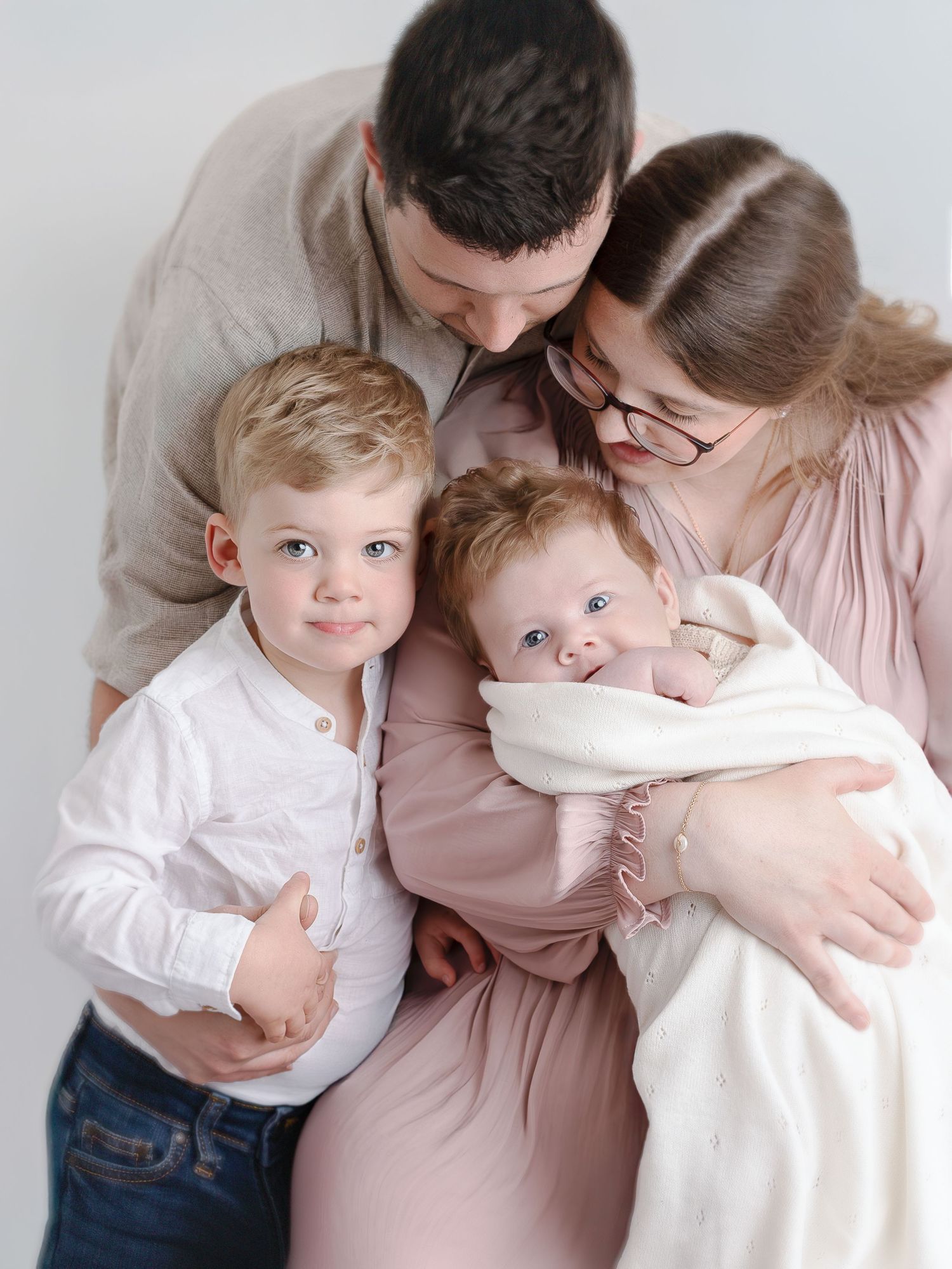 A family of four poses tenderly, parents kissing toddler and newborn baby wrapped in white blanket in a bright studio.