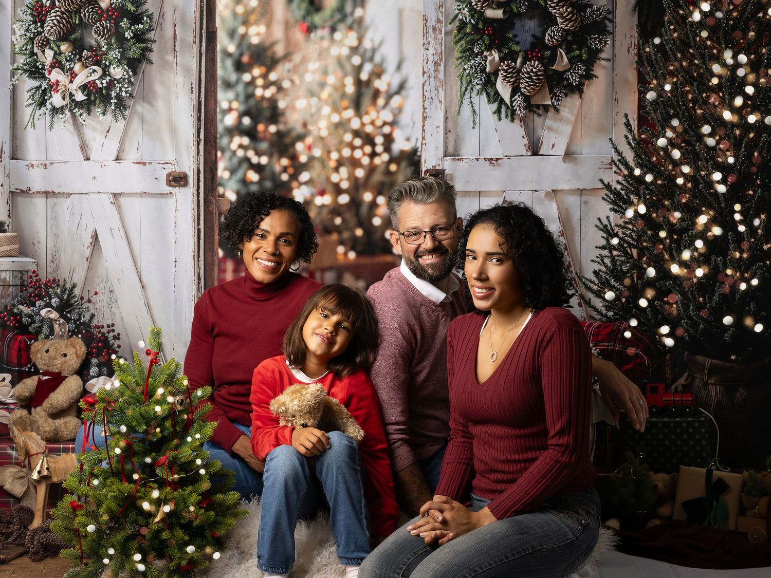 Family of four posing together for a festive Christmas portrait surrounded by decorated trees and holiday wreaths.