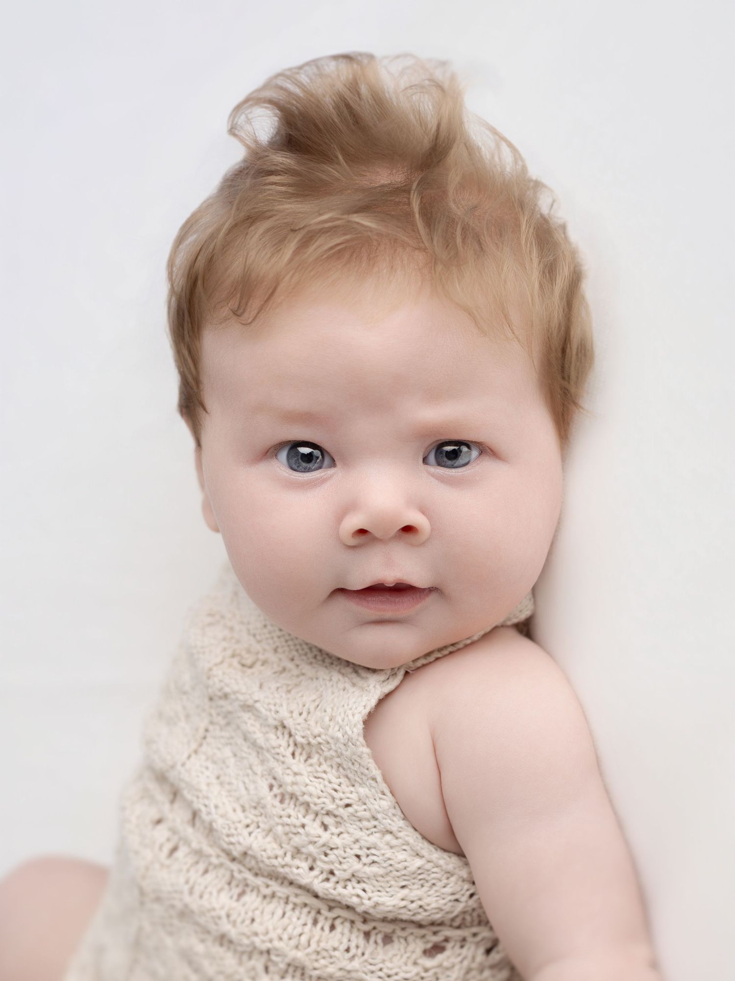 Adorable baby with blonde hair and blue eyes wearing a cream knit outfit, posed against a white background.