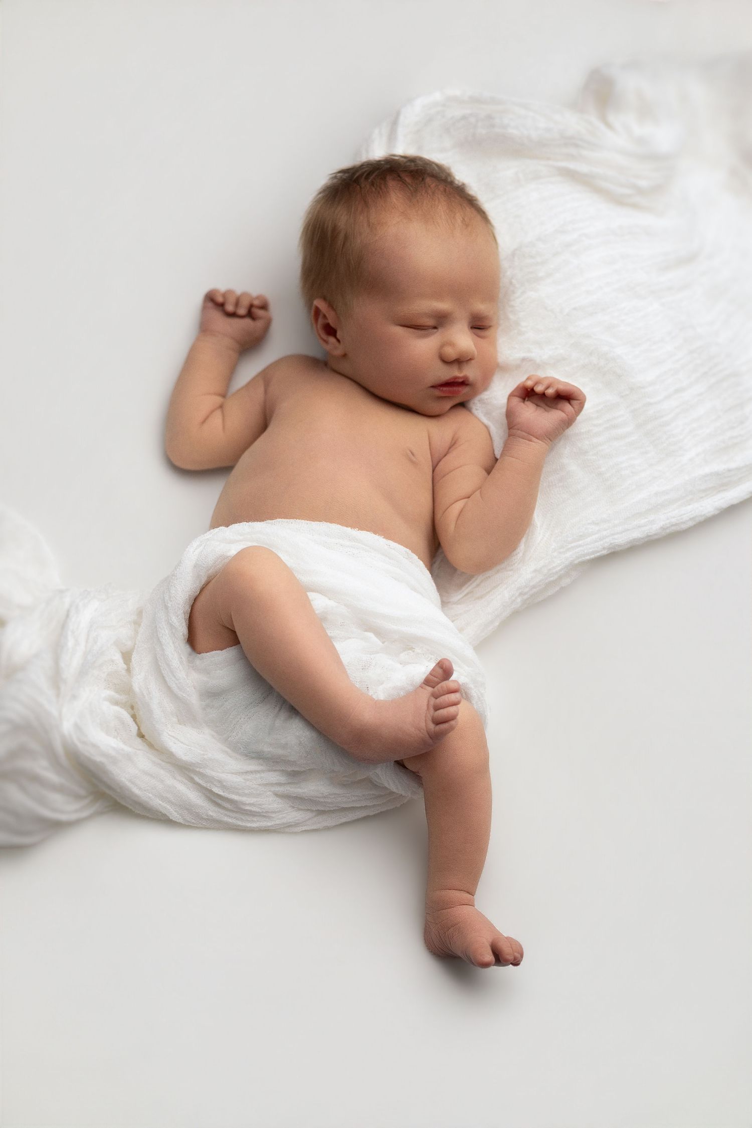 Peaceful newborn baby sleeping on white blanket, wrapped in soft white muslin cloth, arms raised naturally.