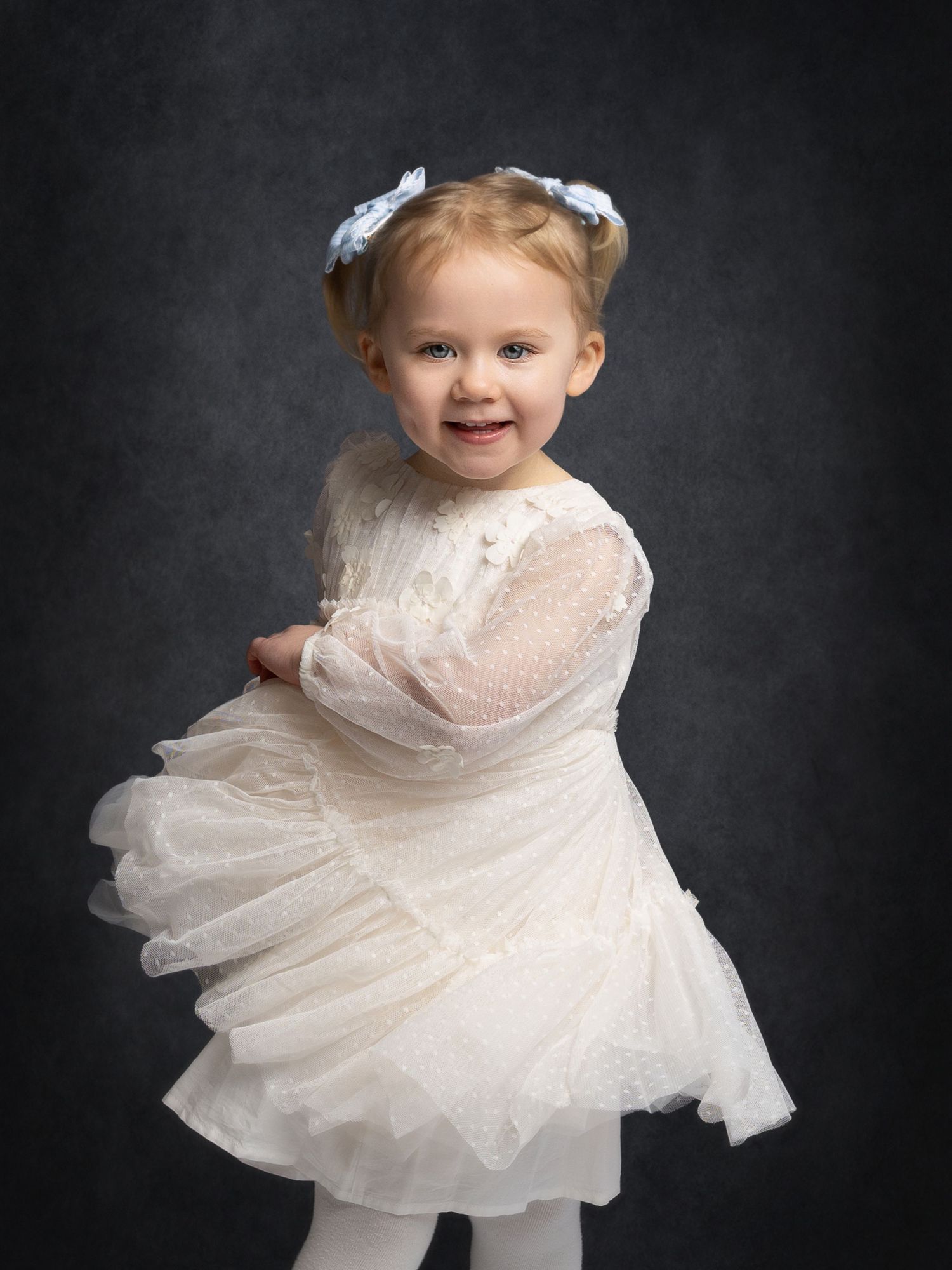 A smiling toddler girl in a white lace dress and blue bow headband poses against a dark studio background.