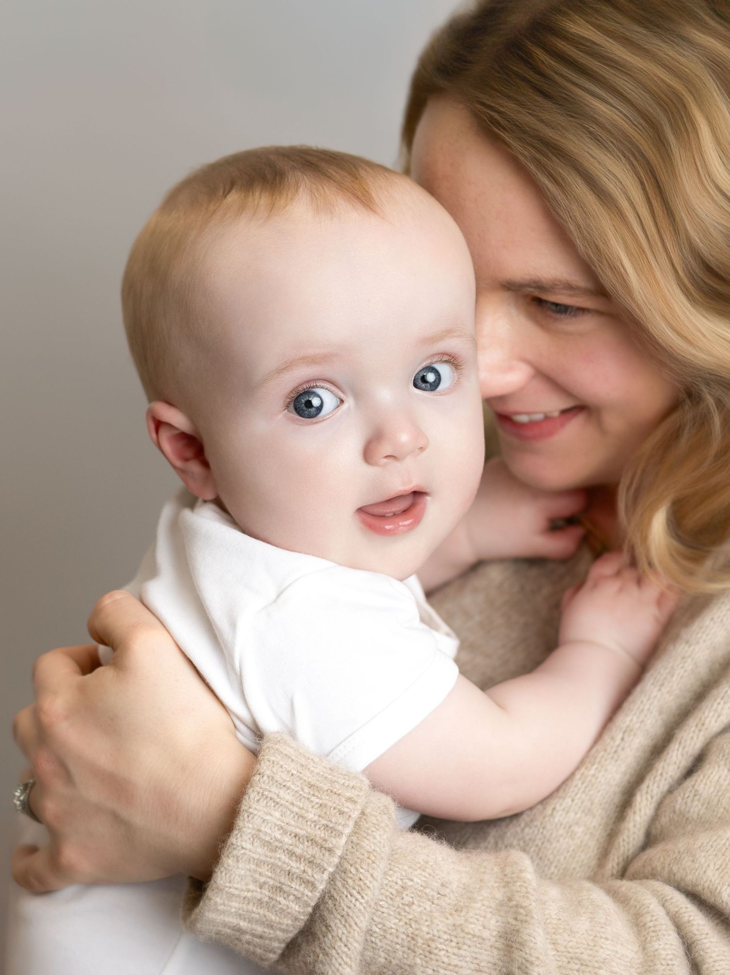 A baby in white clothing looks at the camera with bright eyes while being held in a loving embrace against a beige sweater.