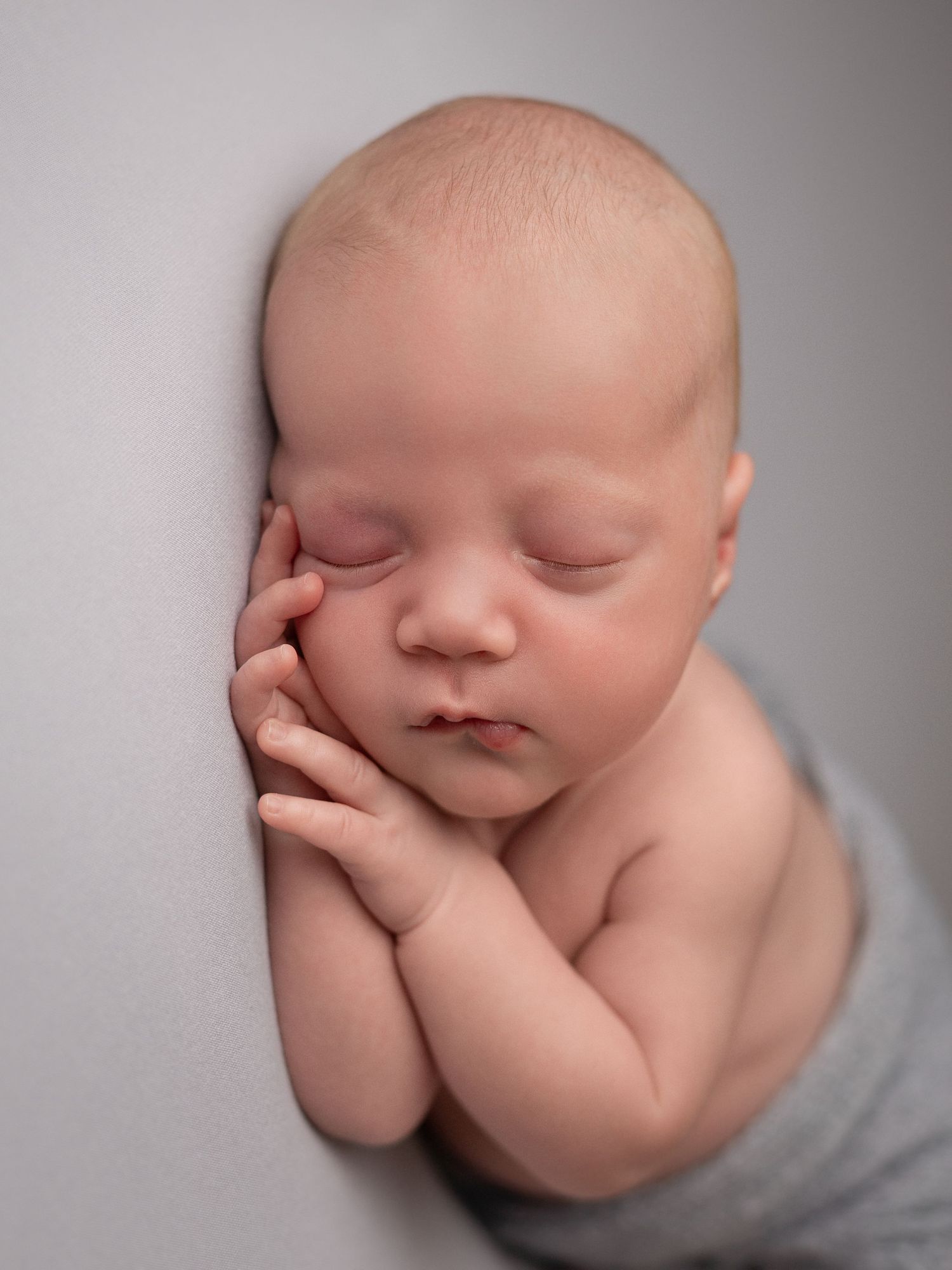 Peaceful sleeping newborn baby with hands tucked under cheek, wrapped in soft gray fabric against neutral background.