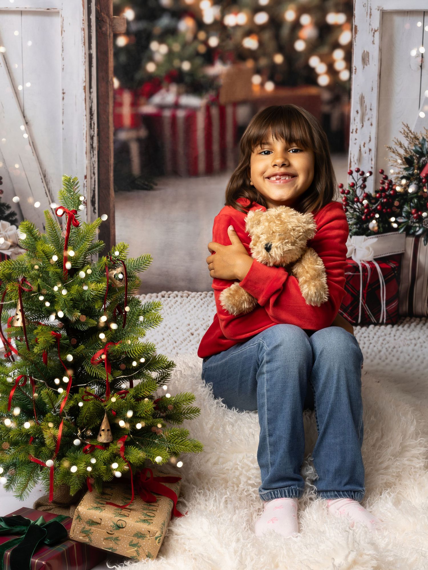 A smiling girl in a red sweater hugs a teddy bear beside a decorated Christmas tree with gifts.