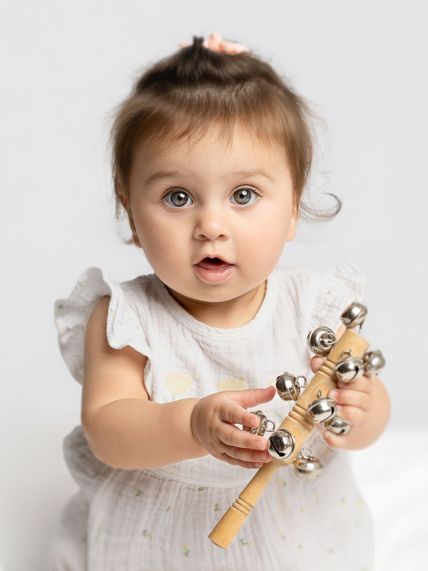 Cute baby girl with big blue eyes holding a wooden bell rattle toy, wearing a white dress with hair up.