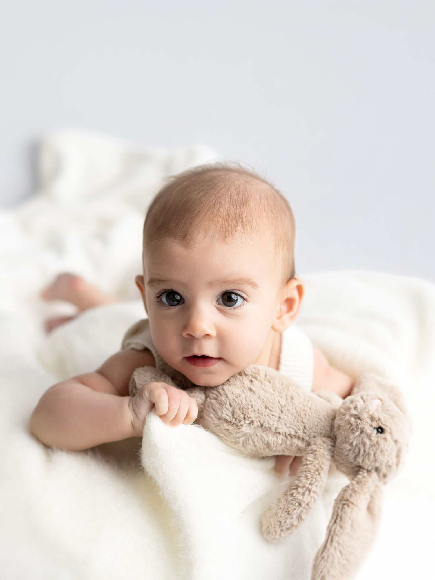 Adorable baby lying on white blanket holding a plush bunny toy, looking curiously at the camera.