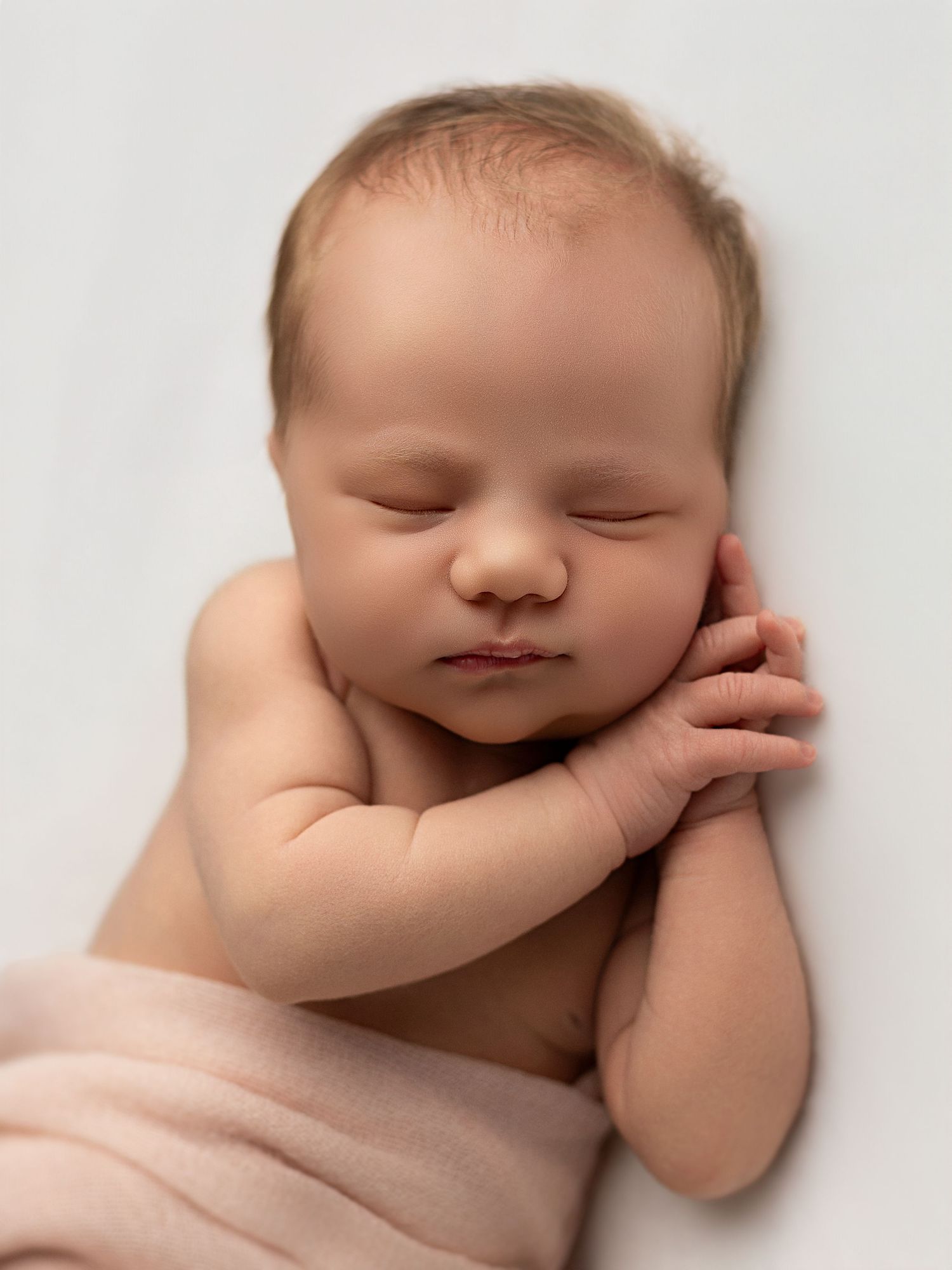 Peaceful newborn baby sleeping on white background wrapped in soft pink blanket with tiny hands near face.