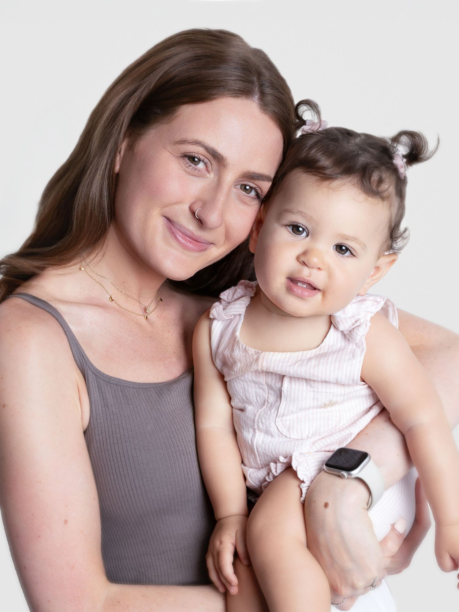A smiling woman in a gray tank top holds a happy baby girl in a pink outfit against a white background.