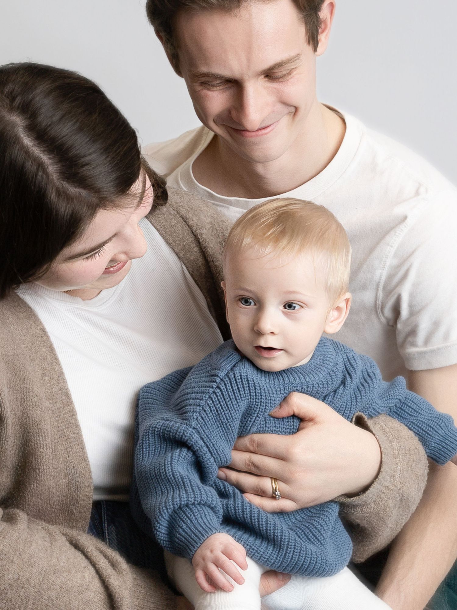 A young family with a smiling man and woman lovingly looking at their blonde baby in a blue knit sweater.