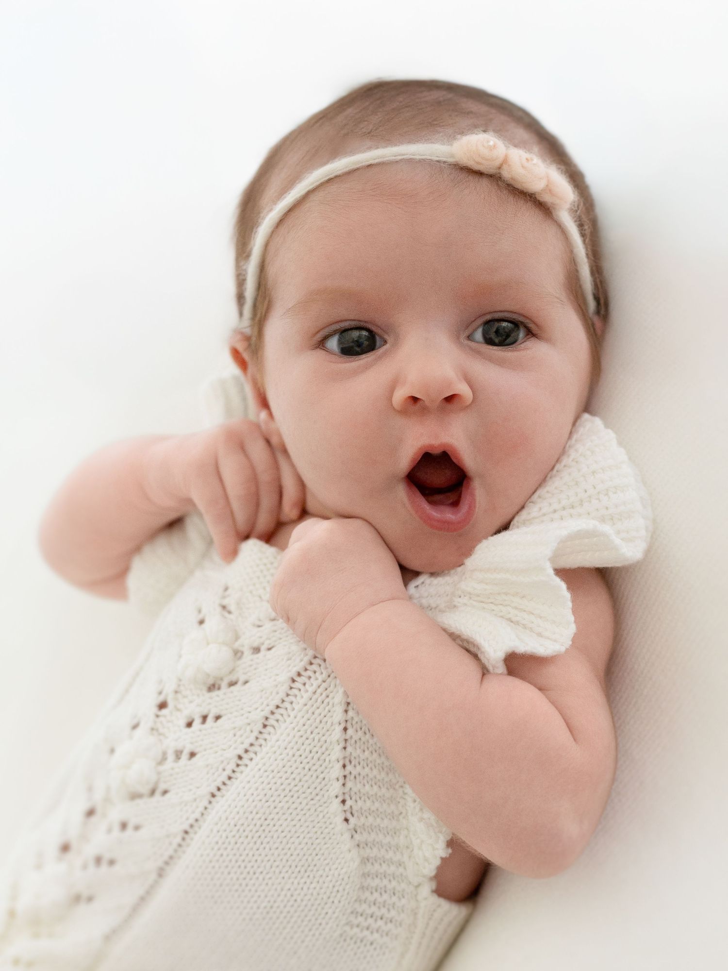 Infant wearing white lace dress smiles and plays in bright studio setting.