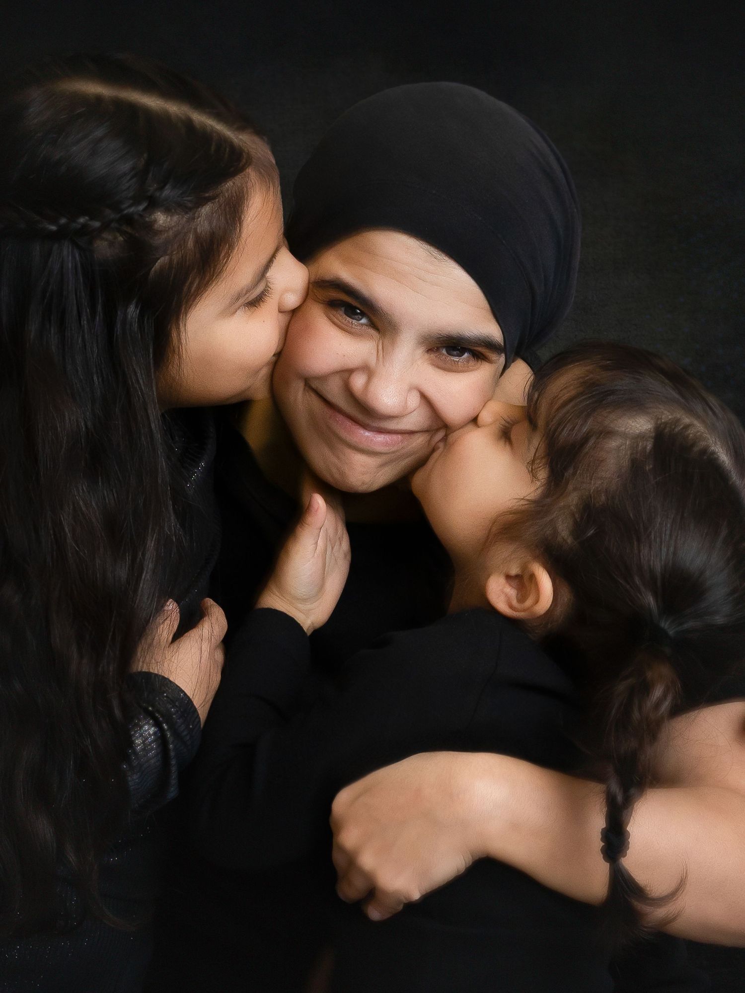 A woman in a black hijab smiles warmly as two young girls kiss her on both cheeks.