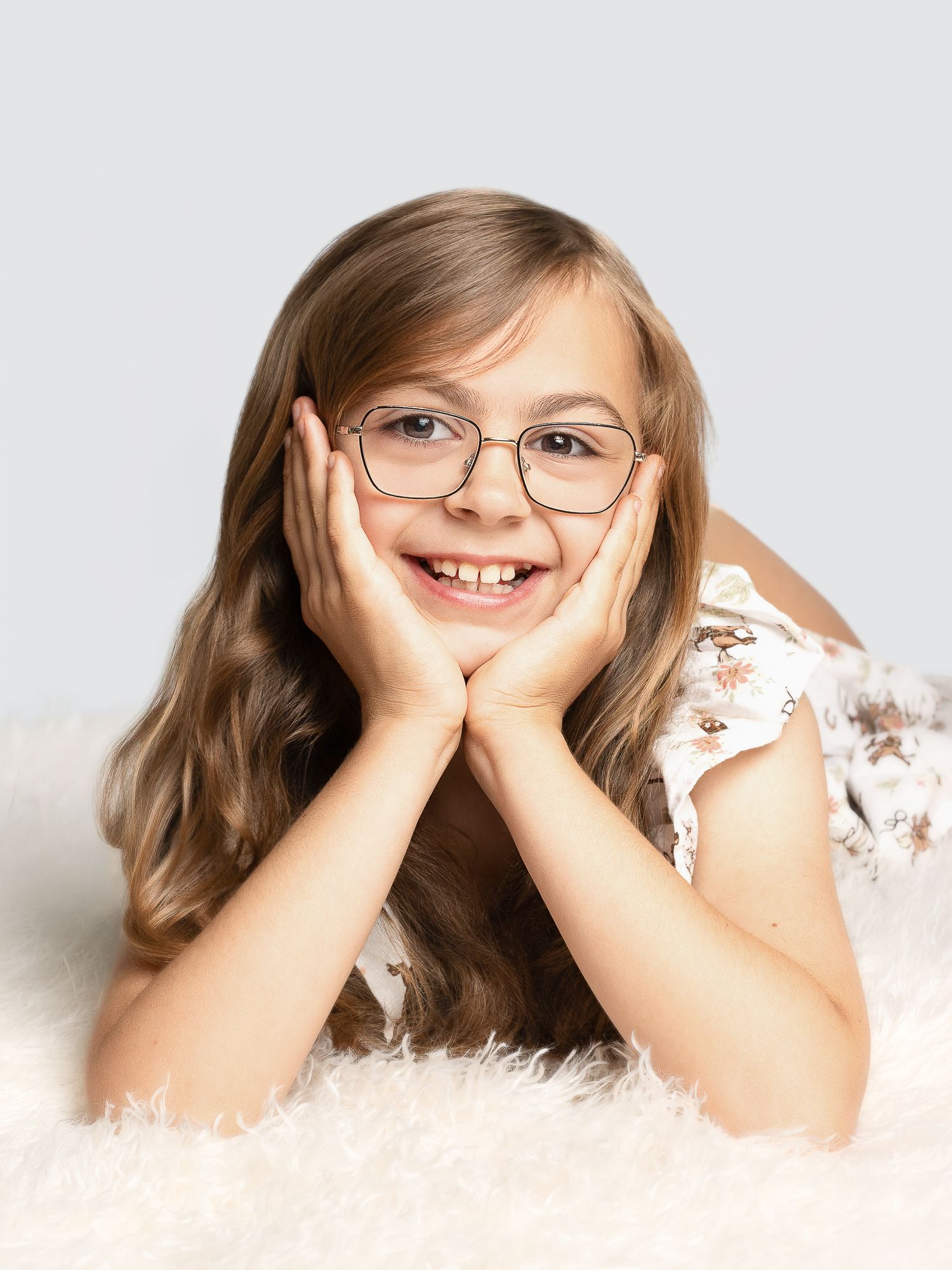 A cheerful young girl with long hair and glasses lying on a fluffy rug, resting her chin on her hands.