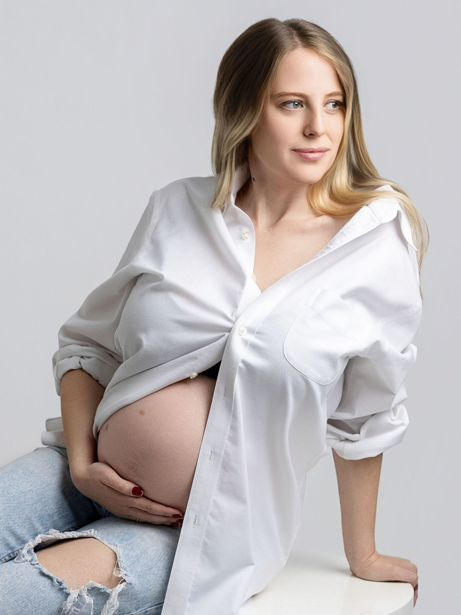 A maternity photoshoot featuring a white button-down shirt and light denim in a minimalist studio setting.