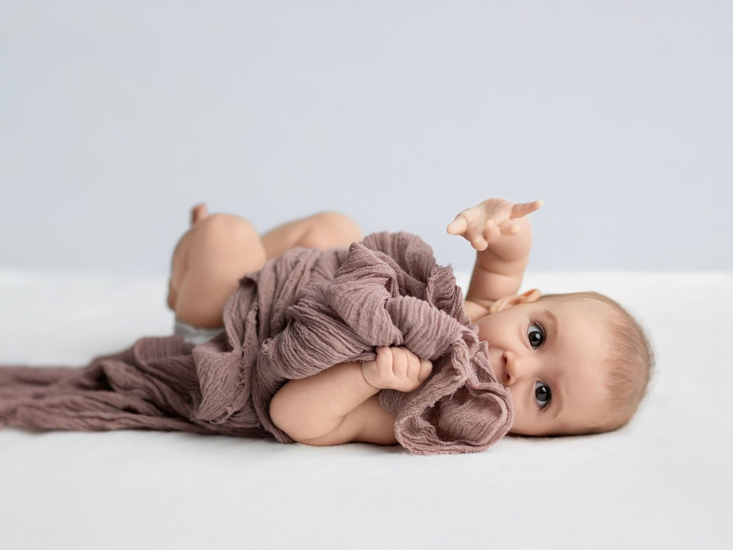 Baby wrapped in mauve fabric lying on light background in artistic poses.