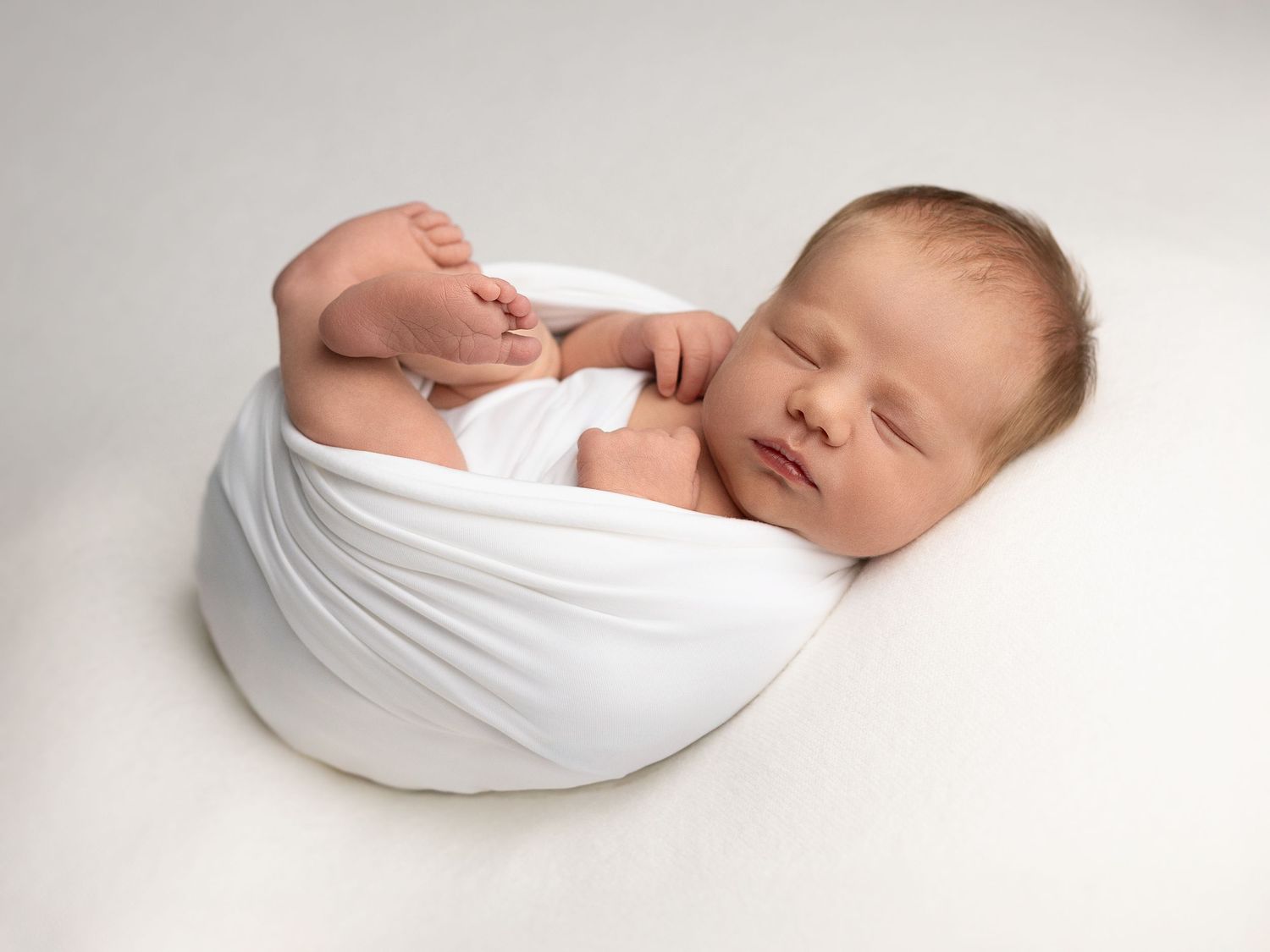 Peaceful newborn photography series with baby wrapped in white fabric against neutral background.