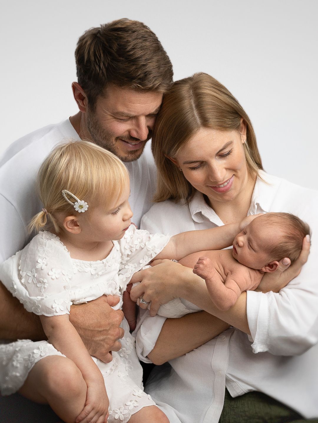 A happy family of four in white outfits, parents smiling at their newborn baby while toddler daughter looks on.