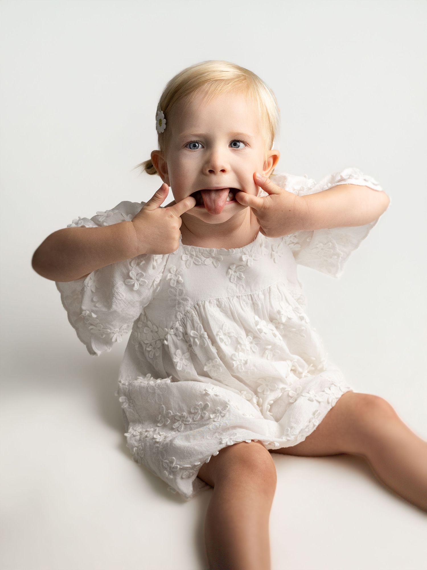 Adorable baby girl in white lace romper making funny faces, sticking tongue out, studio portrait photo.