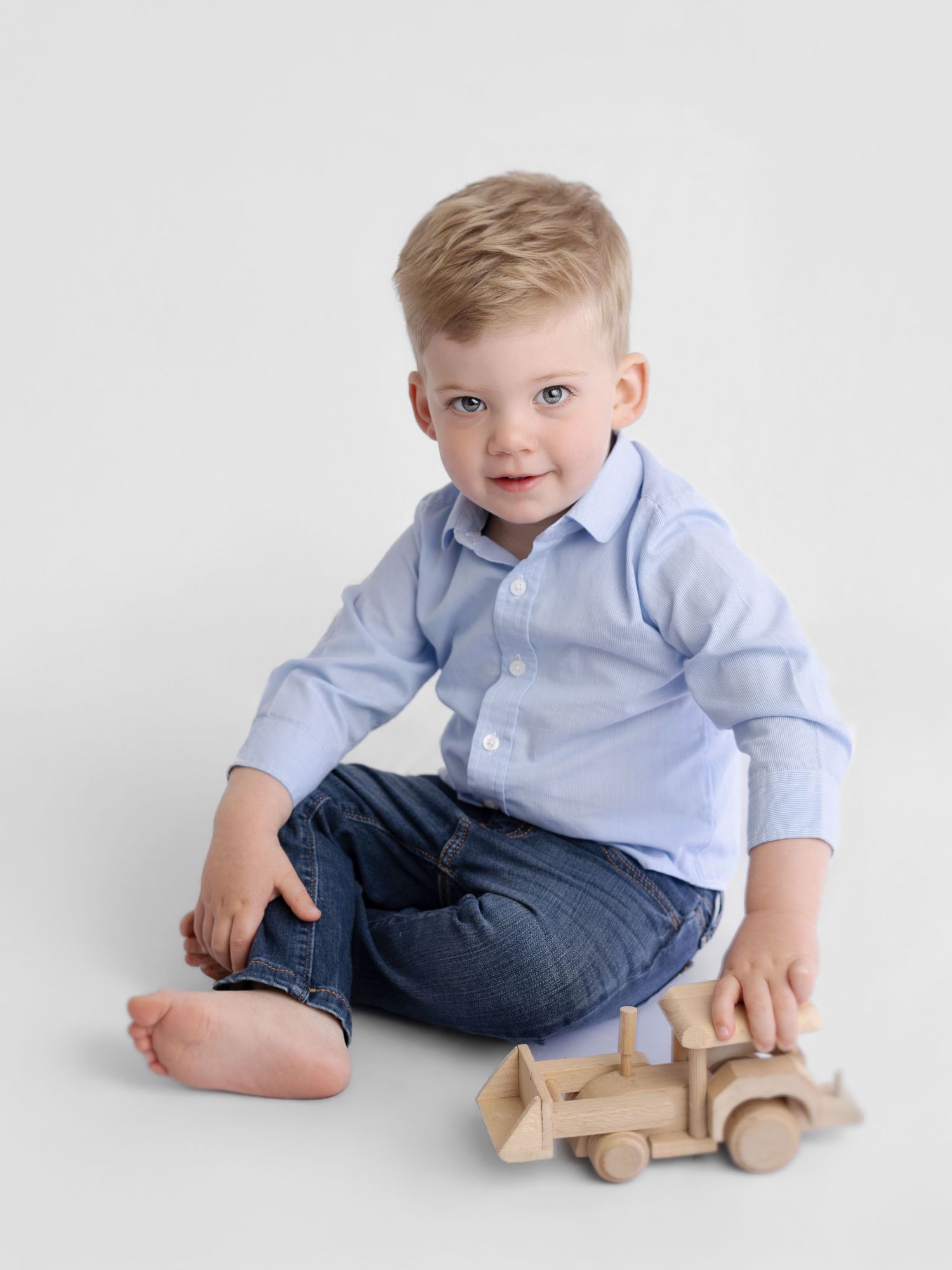 Toddler boy in blue shirt and jeans sits on white background playing with a wooden toy truck.