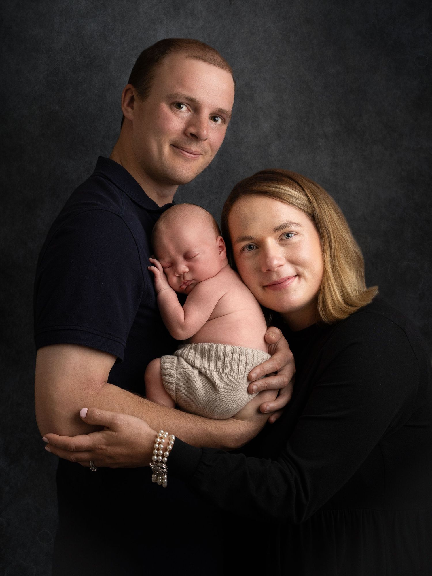 A couple tenderly holds their sleeping newborn baby in a professional studio portrait against a dark gray background.