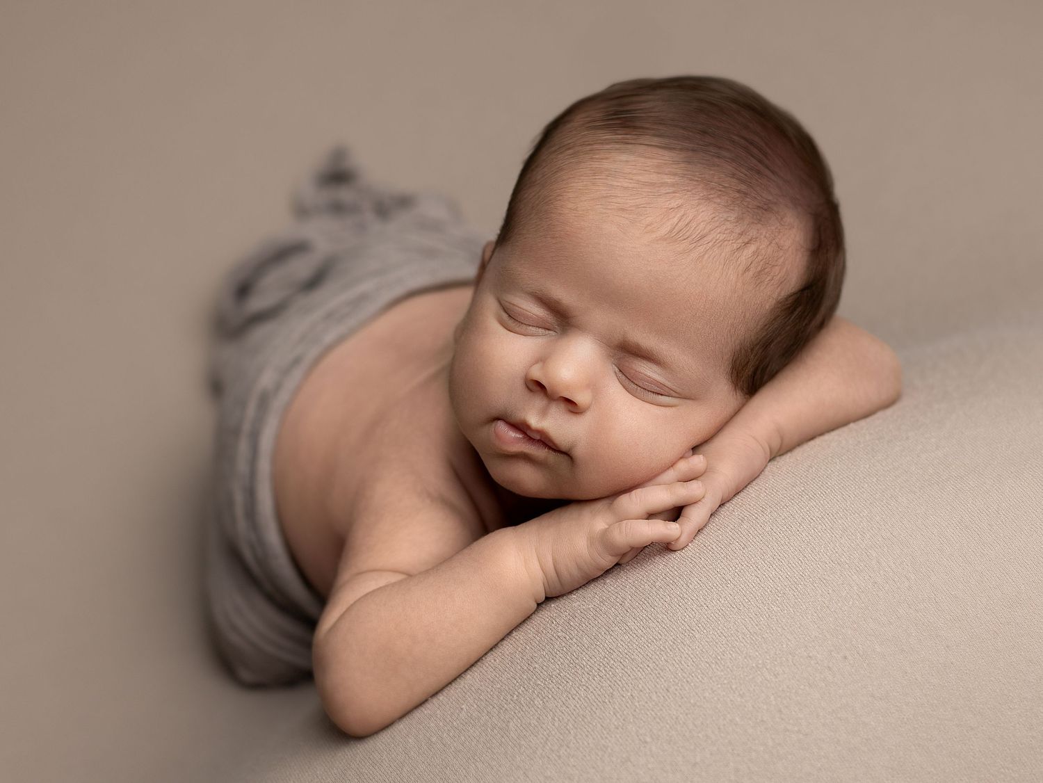 A sleeping newborn baby wearing grey shorts lies peacefully on a beige blanket in a professional studio setting.