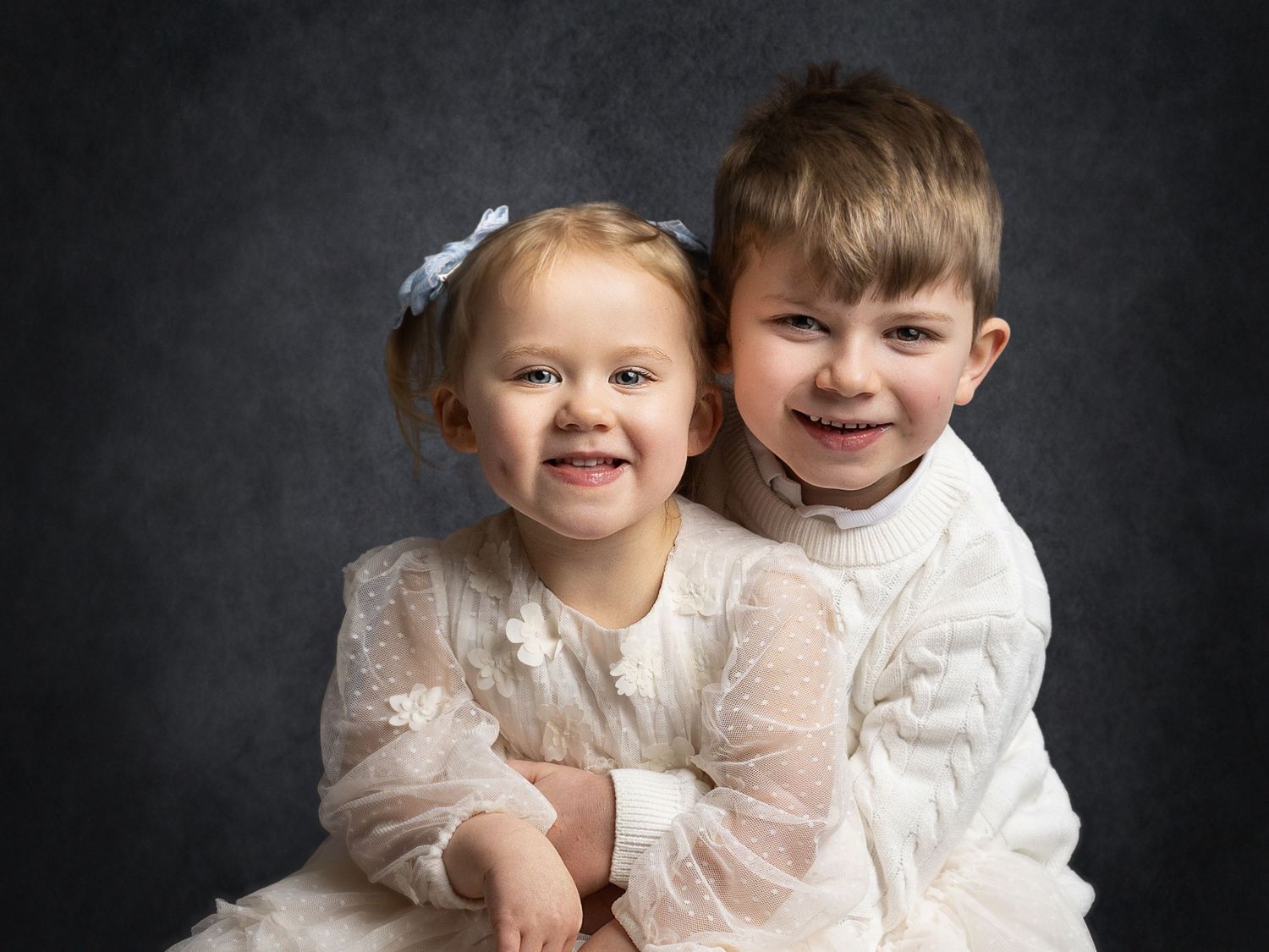 Two young siblings in white outfits smile warmly against a dark gray backdrop in a professional portrait photo.