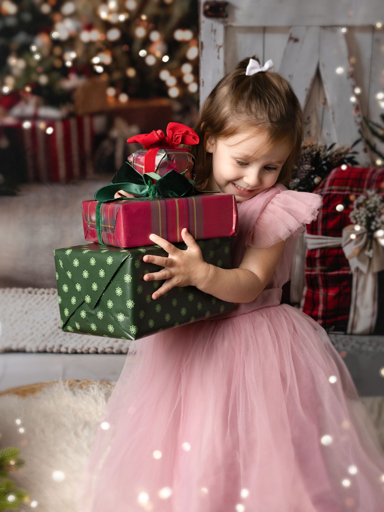 A young girl in a pink tulle dress hugs wrapped Christmas gifts near a decorated holiday tree.