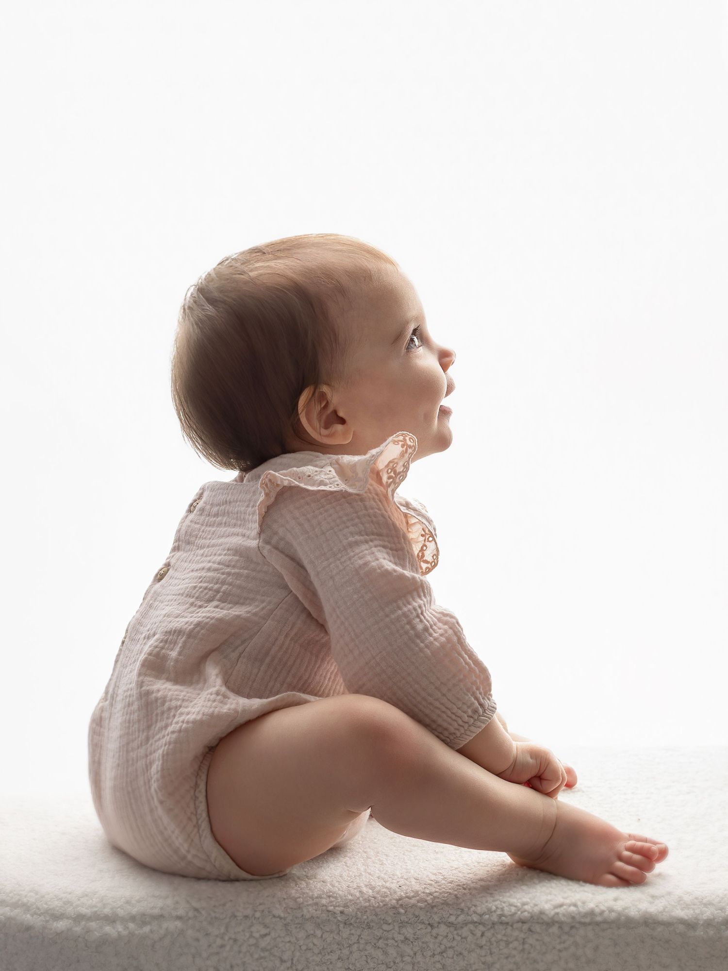 A baby in a pink romper sits on a white surface, gazing upward against a bright background.