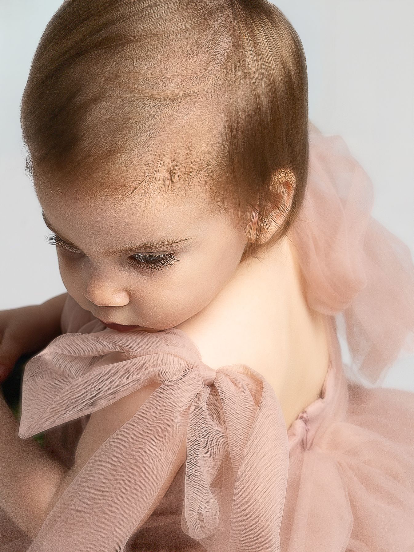 A baby in a soft pink tulle dress with a bow, looking down with delicate features against a light background.
