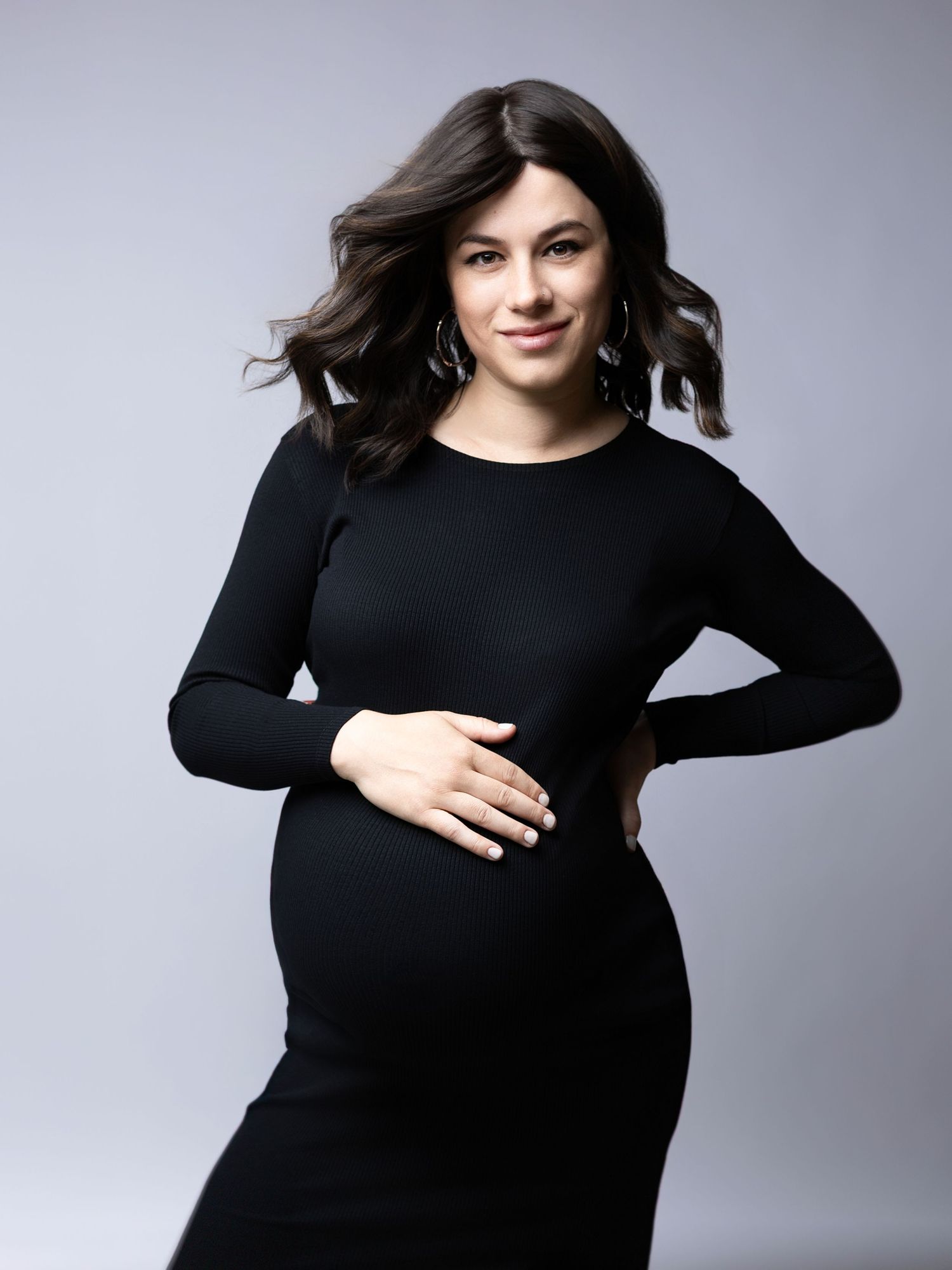 Elegant maternity portrait of woman in fitted black dress against gray background.