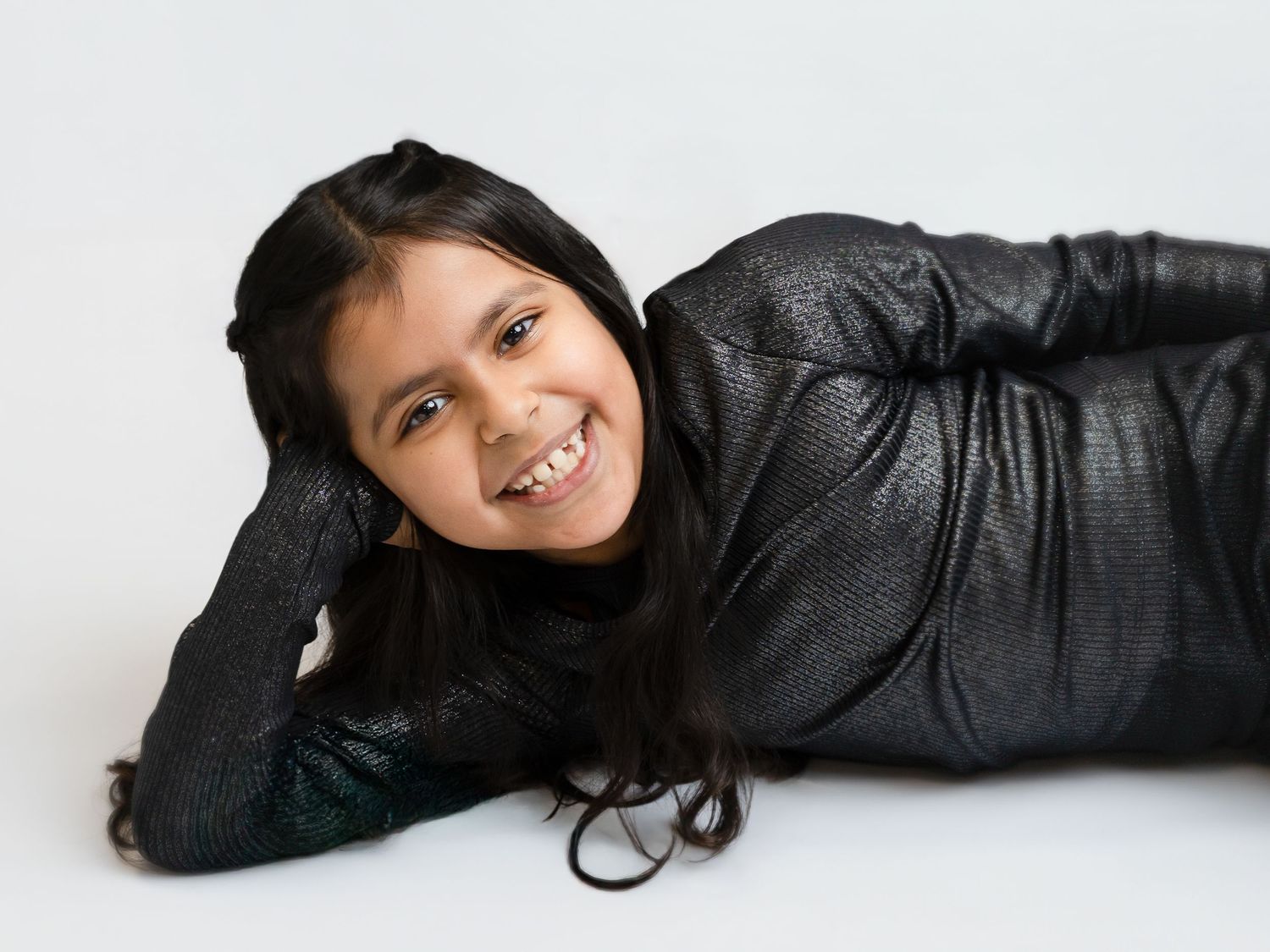 Smiling young girl lying down in a black metallic outfit against a white background.