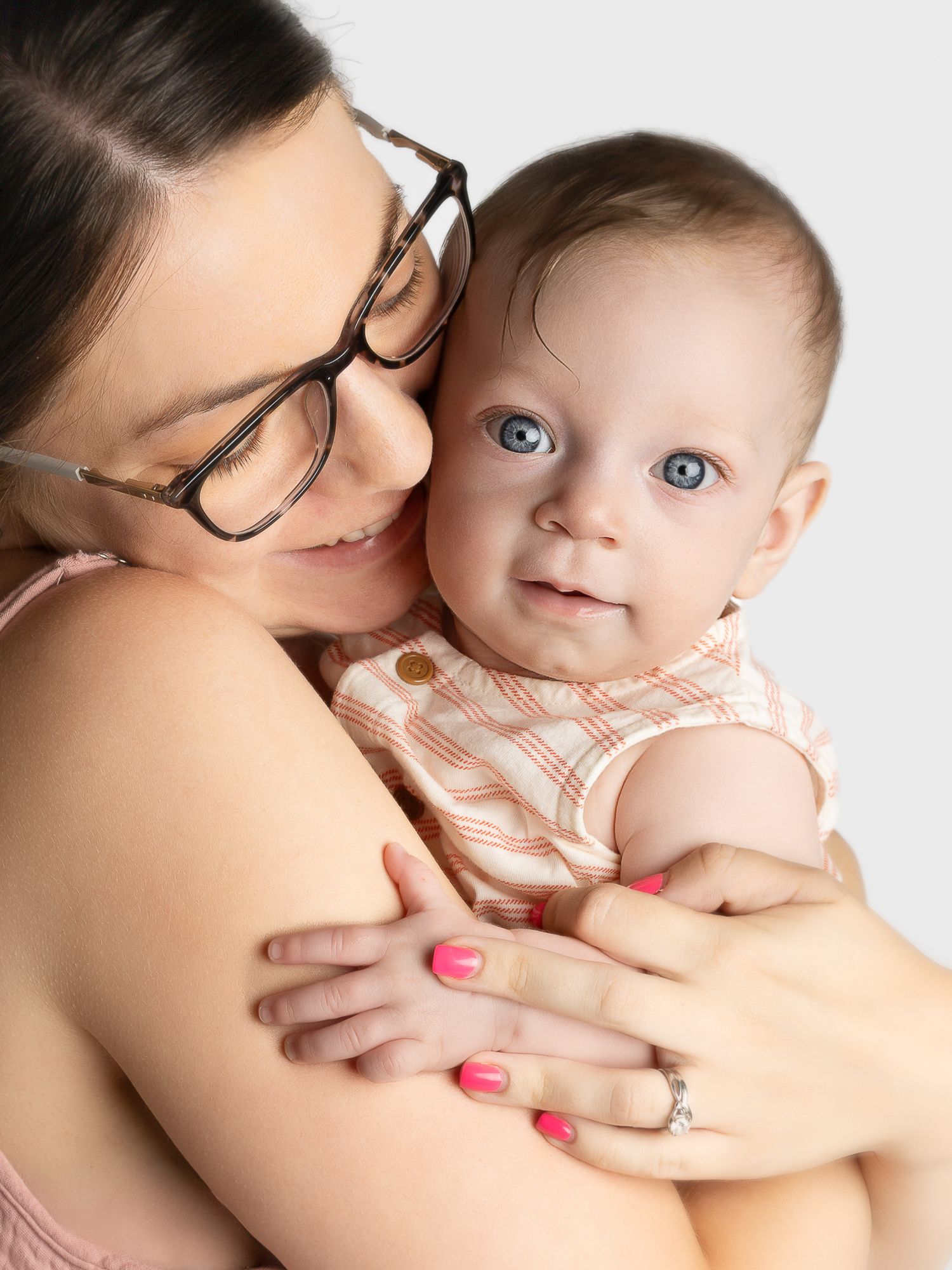 A young mother with glasses and pink nails tenderly holds her blue-eyed baby in a plaid romper against a white background.