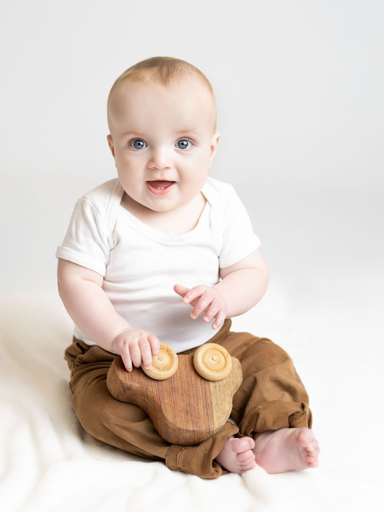 Smiling baby with blue eyes in white onesie and brown pants holds a wooden toy while sitting on a white background.