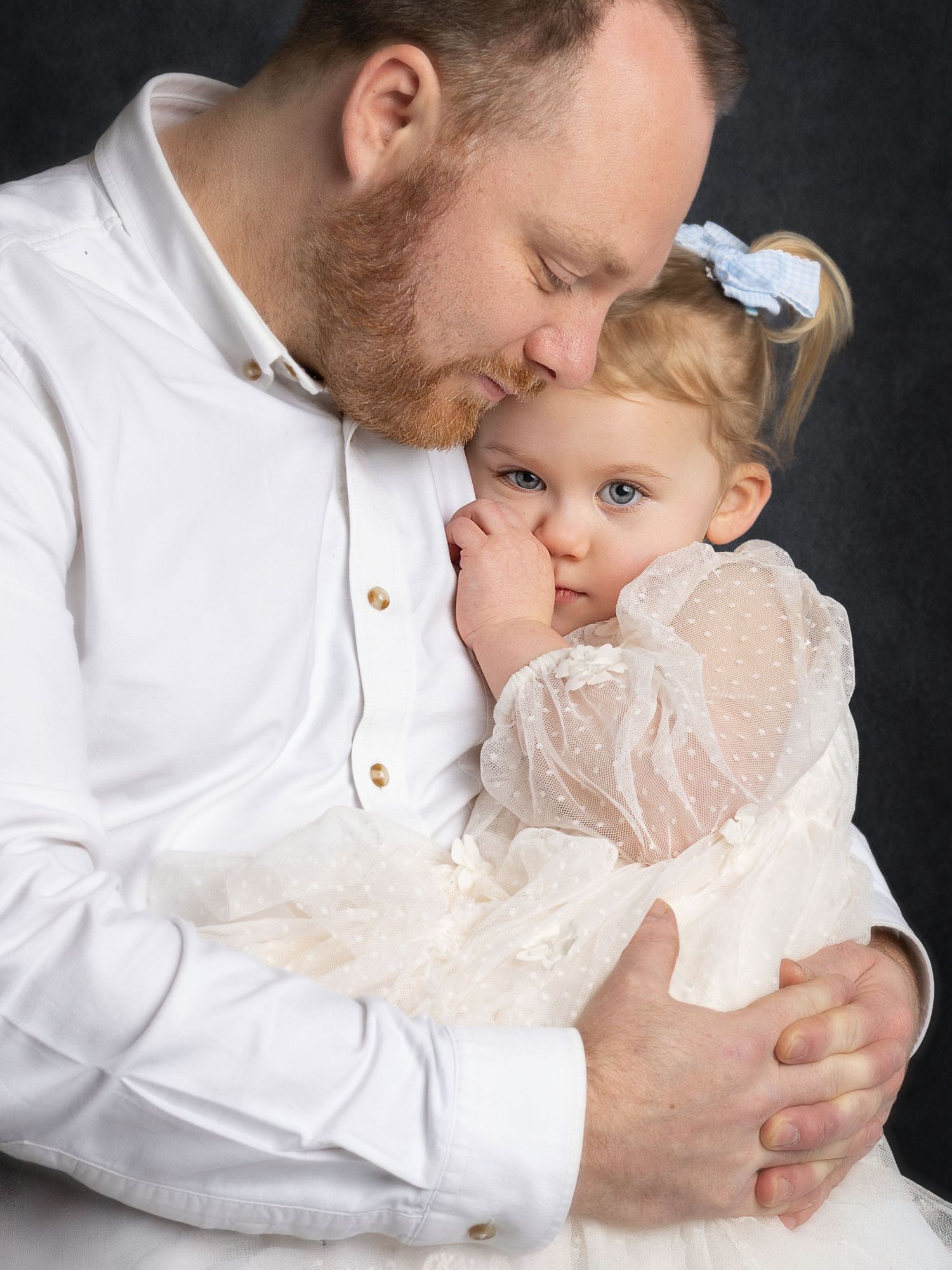 A tender moment as a bearded father in white shirt embraces his young blonde daughter in a lace dress.