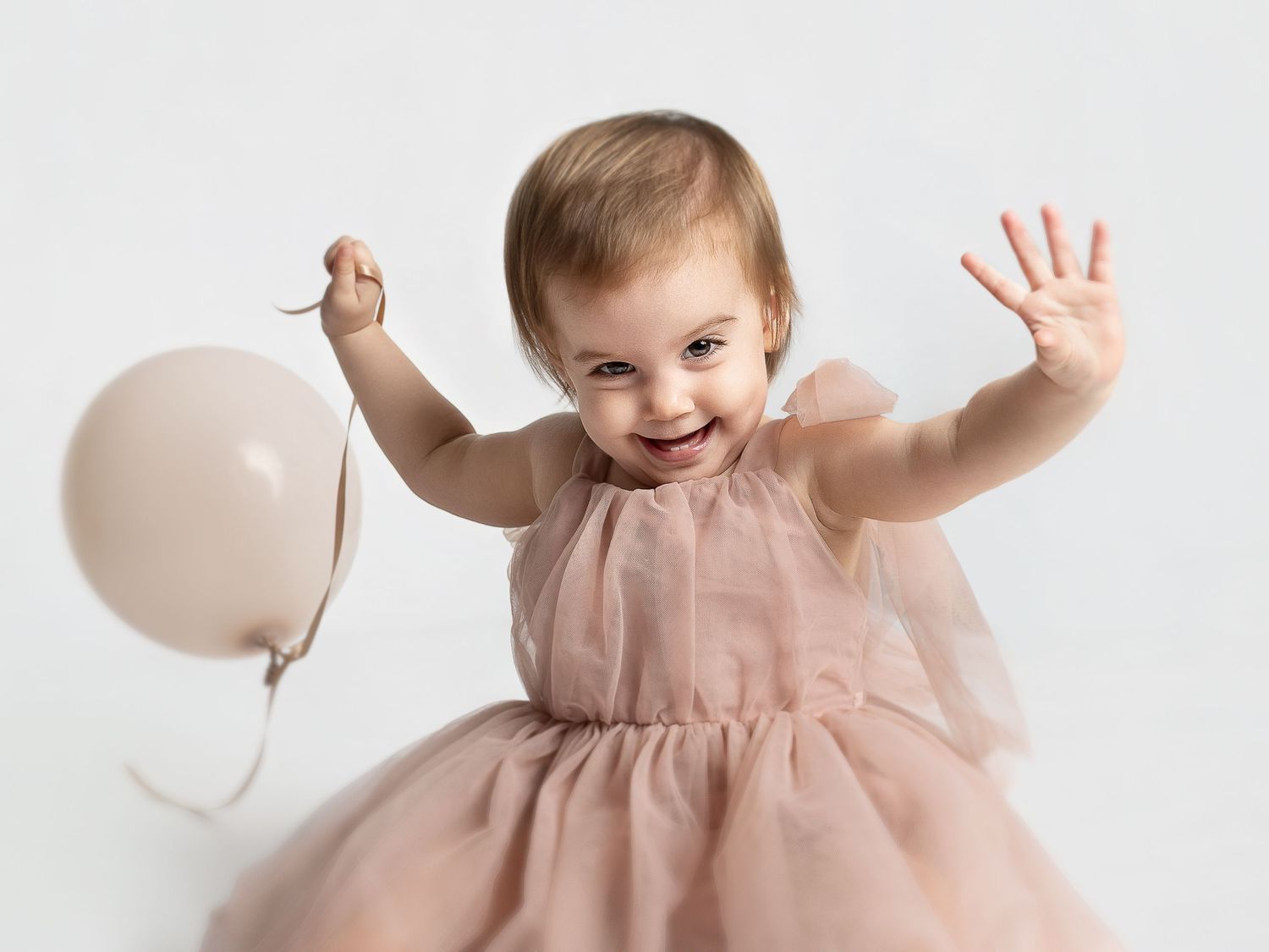 Happy toddler girl in pink tulle dress holding a balloon with arms raised, smiling joyfully against white background.